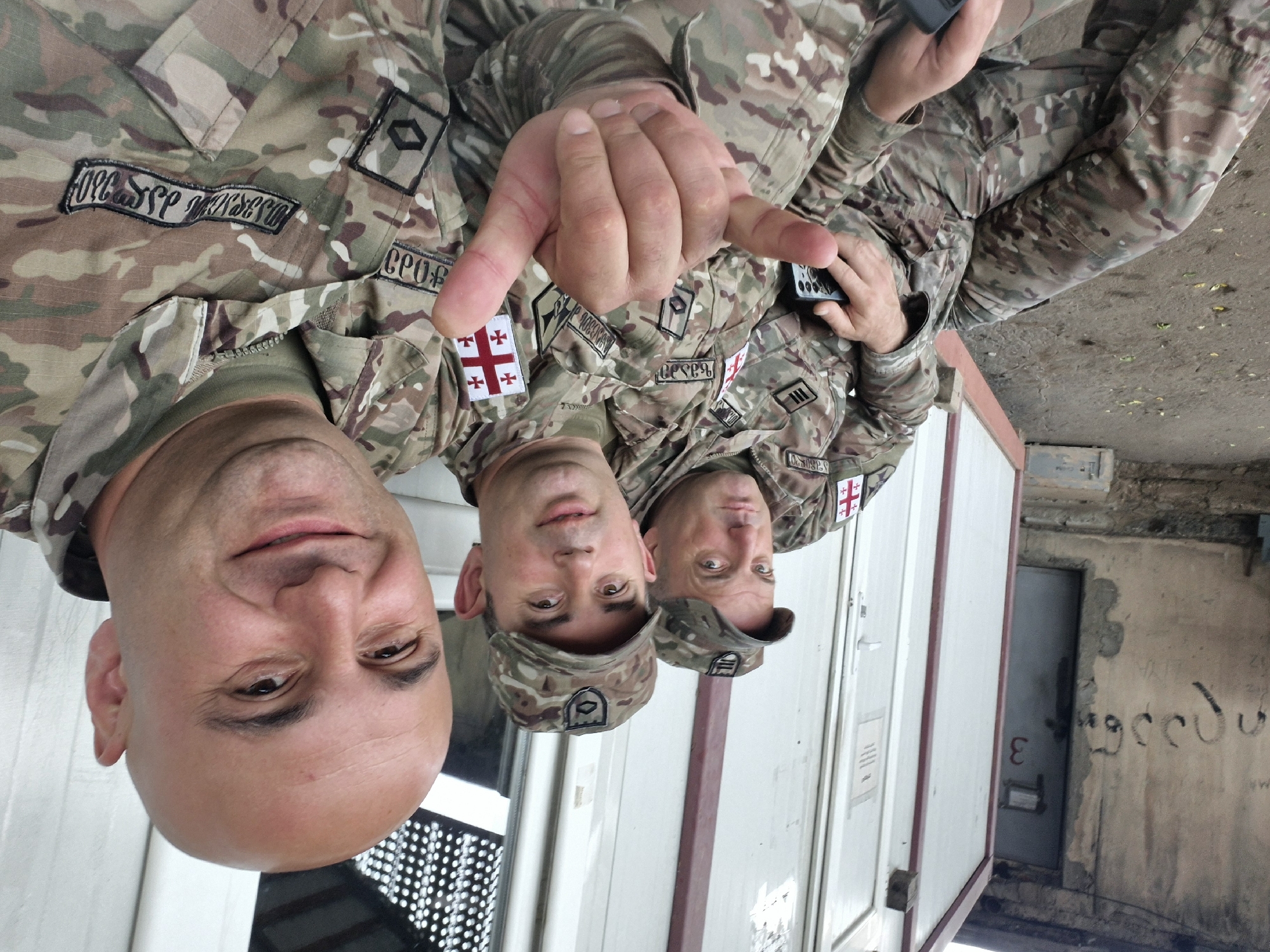 Three men in camouflage uniforms stand in front of a white and red building with a door labeled "3." All three are wearing Georgian flag patches. The wall near the building is concrete and marked with graffiti. The location is likely Tbilisi, Georgia, given the flags.