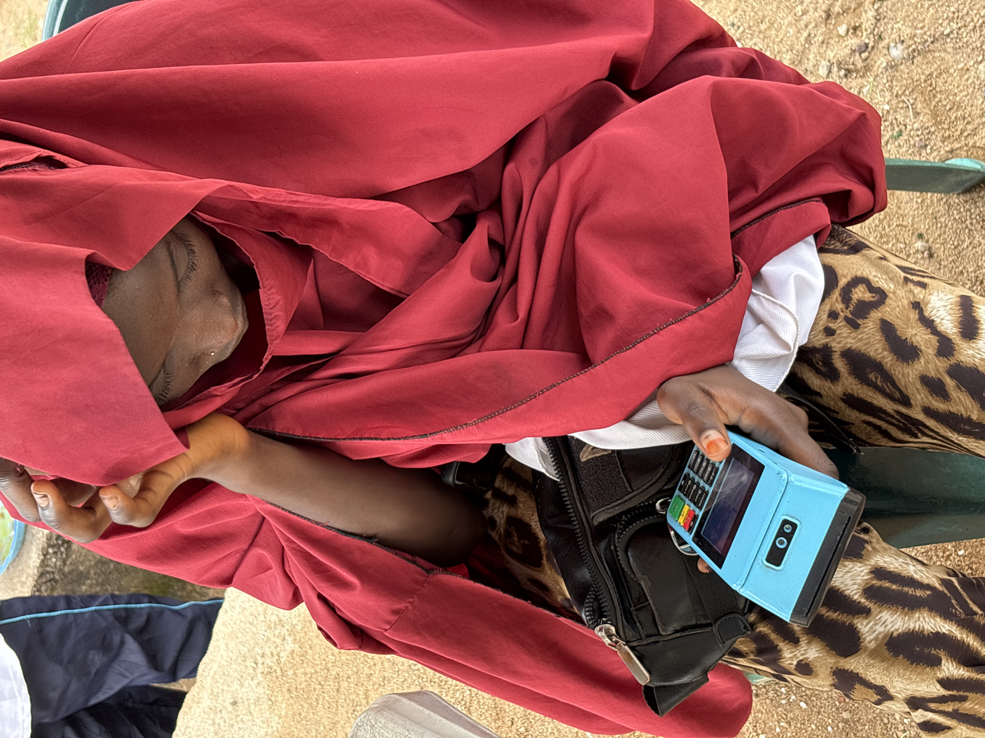 A person in Jos, Nigeria, sits outdoors, dressed in a red hijab and leopard-print pants. They hold a blue credit card machine in their hand, and a black fanny pack is visible. The person appears to be looking down at the machine. The ground is sandy and there is a hint of a person wearing blue trousers in the background.