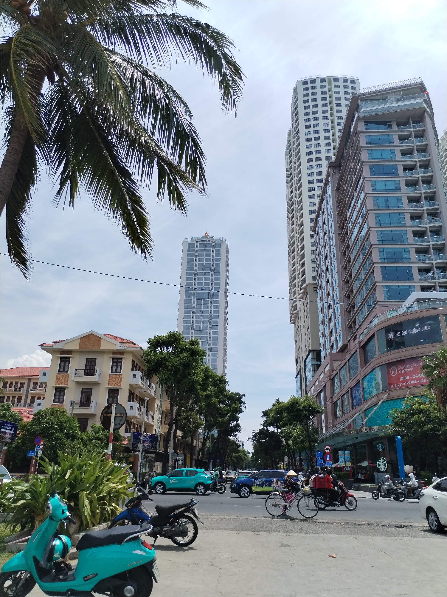 In Nha Trang Ward, Vietnam, a bright, sunny day features a street scene with multiple buildings and various forms of transportation. A tall, slender skyscraper dominates the background, its white facade punctuated by numerous windows. To its right, a wider, multi-story building with a tan and brown exterior and balconies is visible, as is another tall, modern building with a glass facade. In the foreground, palm fronds from a large palm tree partially obscure the sky and the upper portions of the buildings.
The street itself is bustling with activity. Several motorcycles and scooters are parked along the sidewalk, including two bright turquoise scooters in the immediate foreground. A turquoise car is driving on the street, alongside other vehicles and a bicycle. People are riding motorcycles and a bicycle, some wearing conical hats, indicating the cultural context. The buildings lining the street include shops and businesses, with visible signage and advertisements. Lush green trees and bushes add to the urban landscape. The sky is partly cloudy, with a bright, clear sky visible through gaps in the clouds.