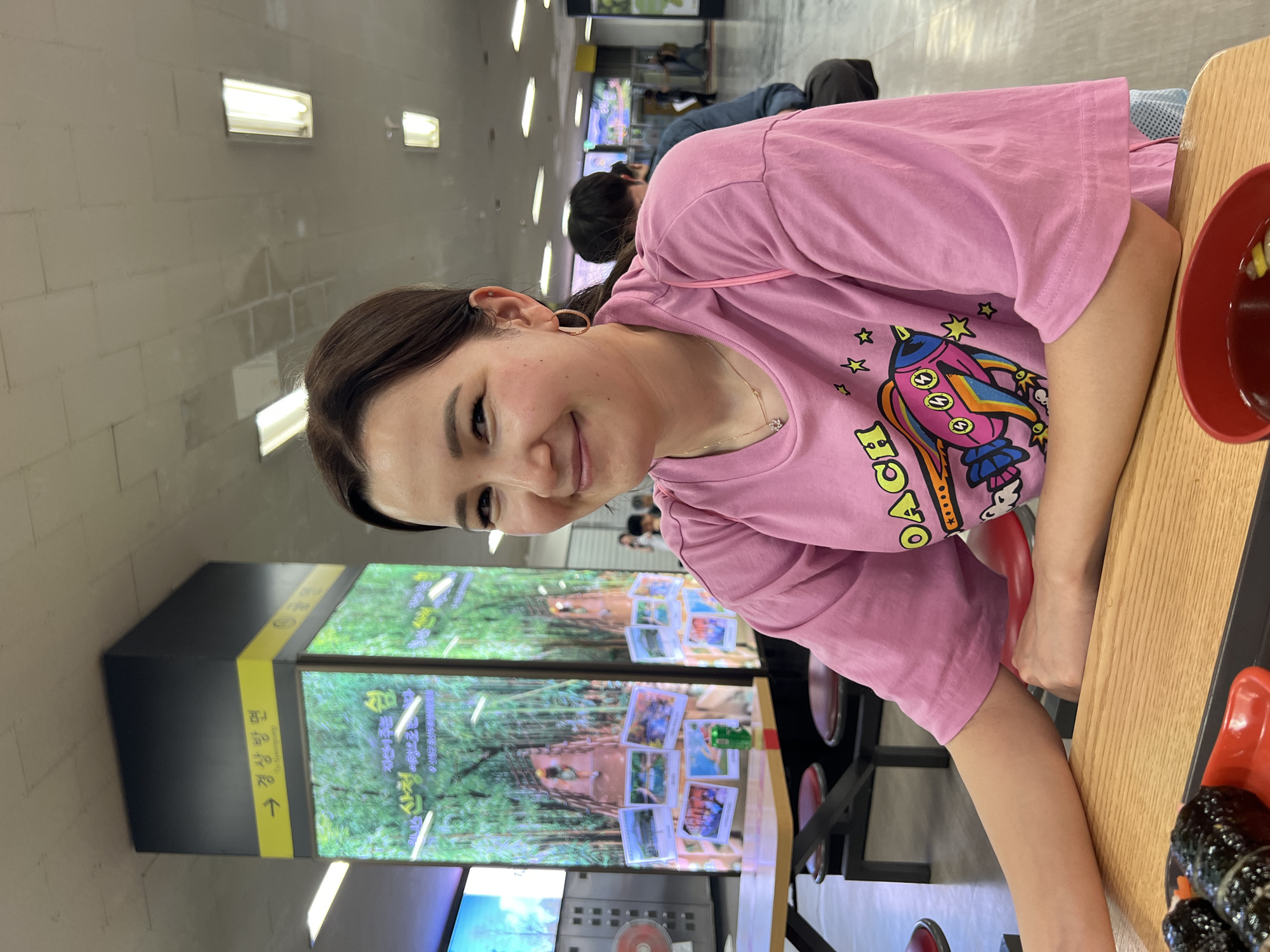 A young woman with dark hair pulled back smiles at the camera, her head tilted slightly to the left. She is wearing a pink t-shirt with a cartoon rocket graphic on it and a delicate necklace. She has on hoop earrings. She is seated at a light wood table in what appears to be a food court or cafeteria. In front of her on the table is a red plate with portions of kimbap, a type of Korean seaweed rice roll. To the right of the plate is a small red bowl containing a dark liquid, possibly a dipping sauce or soup. Behind her, a large column is decorated with a digital display showcasing images of a lush green forest path and several small photographs. A yellow sign on the column with Korean text and an arrow indicates directions. Other patrons are visible in the background, some seated and others standing. The ceiling features fluorescent lighting, and the overall environment suggests an indoor public space.