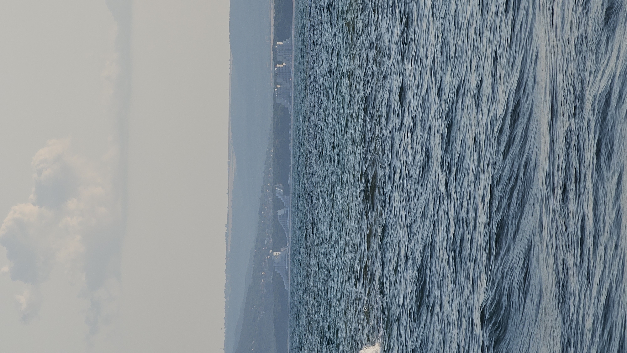 This media file captures a scenic view of the Black Sea coast, likely near Balchik, Bulgaria. The image is oriented vertically, dividing the frame into two distinct halves. On the left, a hazy sky dominates, with large, soft white clouds appearing to drift across a muted, pale background. The haziness suggests either a smoky atmosphere, possibly from distant fires, or a misty morning.

The right half of the image is filled with the deep blue, textured surface of the sea. The water is in motion, with visible waves and ripples creating a sense of movement and depth. The light plays across the surface, highlighting the crests and troughs of the waves.

In the distance, along the horizon where the sky meets the land, a coastline is visible. This area appears to be a developed settlement, with several buildings, likely hotels or apartment complexes, dotted along the shore. Behind the buildings, a gentle slope of land covered in dark green vegetation rises, forming a backdrop to the coastal town. The overall impression is one of a serene, perhaps slightly overcast, day at the seaside. The time of day appears to be morning or late afternoon, given the soft, diffused light. No text is visible in the image.
