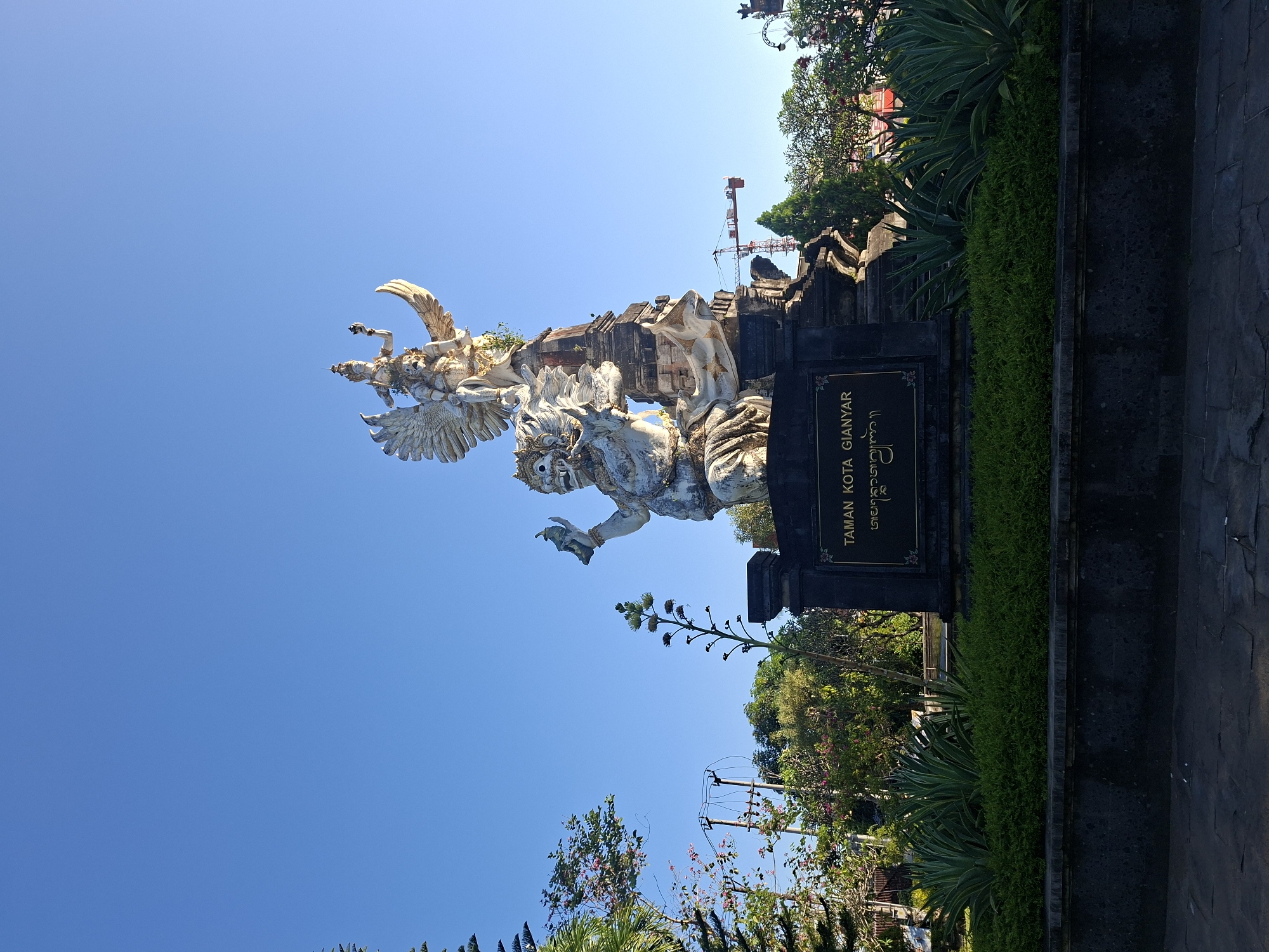 This is a low-angle, eye-level, outdoor, full shot of a large, white stone statue located in the city of Gianyar, Indonesia, under a clear, bright blue sky. The statue depicts a mythological scene with a large, imposing figure at the bottom, possibly a demon or rakshasa, holding an object in its right hand. Above this figure, a smaller, more graceful figure, likely a deity or a prince, is seated on its shoulders, with large, spread wings. The statue is mounted on a dark stone pedestal with a plaque that reads "TAMAN KOTA GIANYAR" in English and a script below that appears to be Balinese. The statue is surrounded by lush green manicured bushes and plants, with some taller plants and trees in the background. To the left, there are some more trees and a utility pole with wires. In the distance, a construction crane is visible. The lighting suggests it is either early morning or late afternoon, with the sun casting a warm glow. The scene appears to be a public park or square, a landmark within the city of Gianyar.