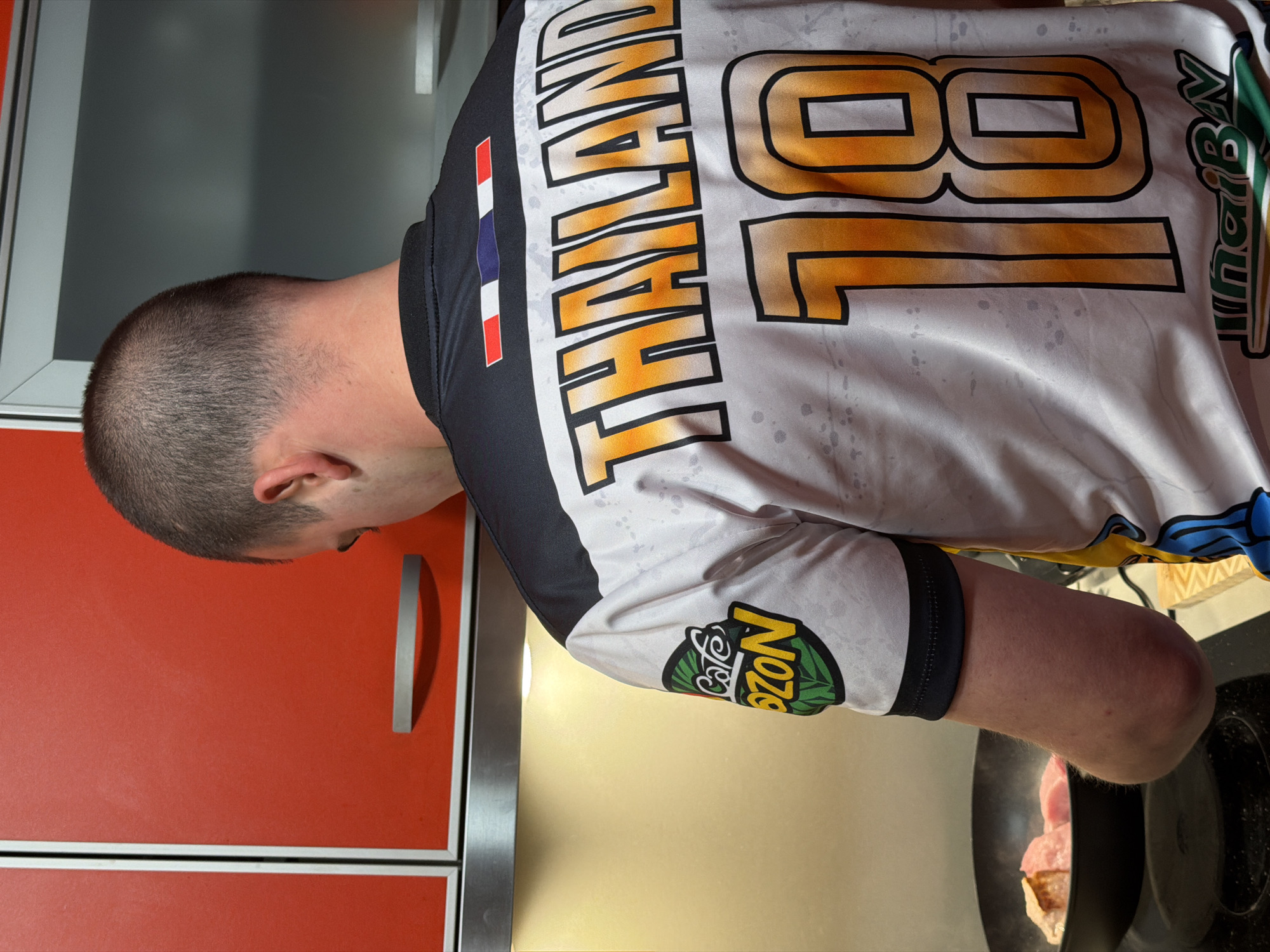 The image, taken in Varna, Bulgaria, shows a person from behind, bending over a cooking pan in what appears to be a modern kitchen. The person, likely male, has short, buzz-cut hair.
The setting features bright orange kitchen cabinets with silver handles on the left side of the frame. On the right, a black cooking pan is on a stovetop, containing pieces of meat (possibly pork or poultry) that are being cooked, indicated by visible steam or smoke.
The person is actively engaged in cooking, leaning forward to observe or tend to the food in the pan.
Notable details include the person's attire: a white, black, and orange athletic-style shirt. The back of the shirt prominently displays the word "THAILAND" in large orange letters outlined in black, with the large number "18" next to it. On the upper left shoulder, a small Thai flag emblem (red, white, blue, white, red stripes) is visible. The right sleeve features a circular logo with a green leaf design and the text "Cafe Dozon". A partially visible text "JunglBv" is also present on the lower right back of the shirt. The lighting suggests an indoor environment, potentially evening or under artificial light. No specific emotions are discernible from the person's posture.