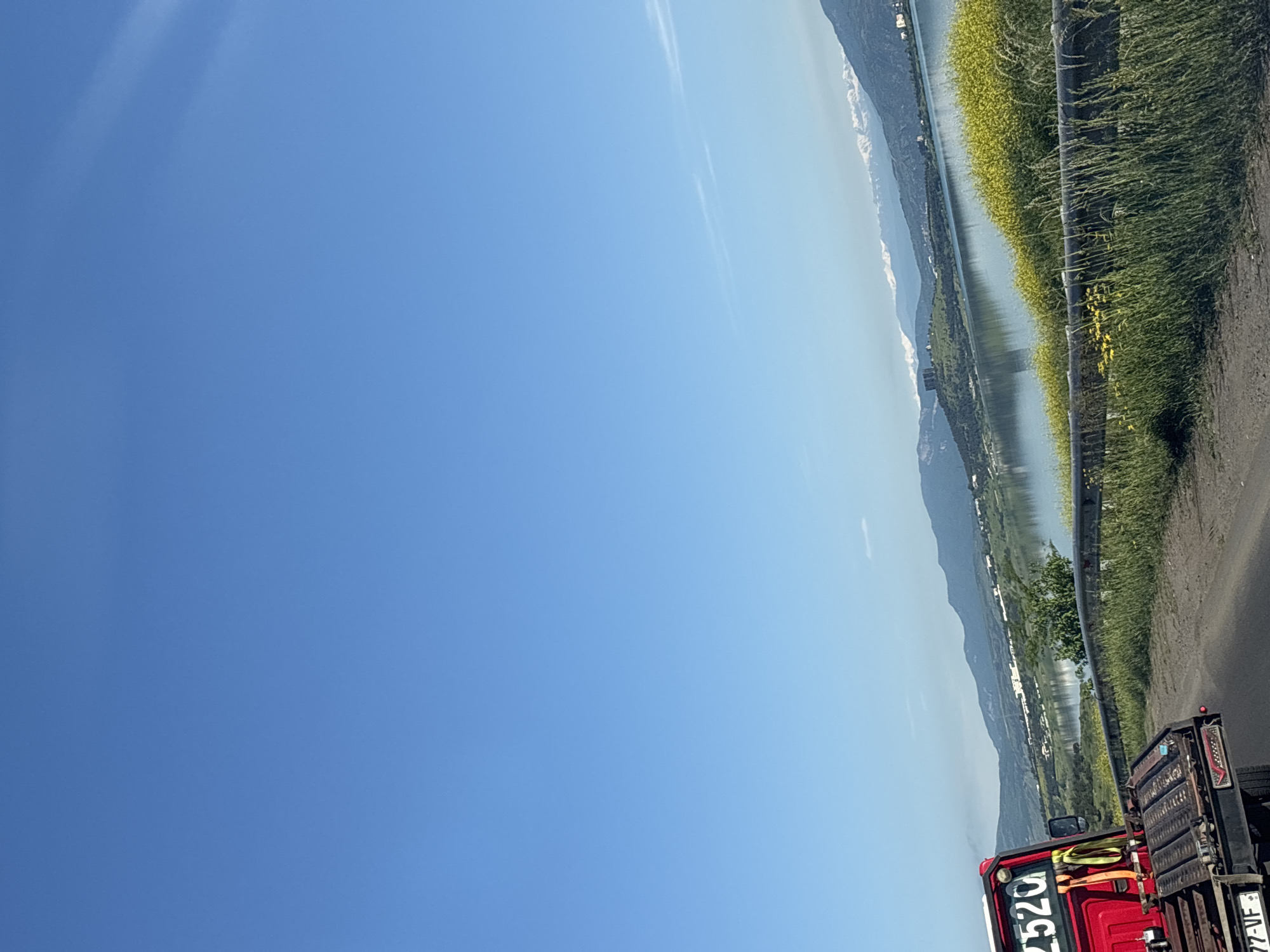 The image shows a view from a vehicle, likely a truck, on a road. The scene includes a body of water, possibly a lake or river, bordered by greenery on the right side of the frame. Mountains are visible in the distance beyond the water. The sky is clear and blue, taking up a large portion of the image. The vehicle's red cab is partially visible in the lower right corner, with "2520" written on the front. Part of the vehicle's rear is also visible, with a partial license plate showing "77-VF". I cannot determine the specific location in Tbilisi, Georgia.