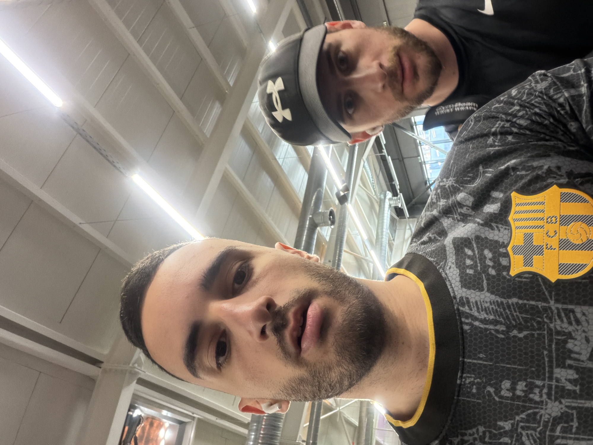Two men are in an indoor setting. The foreground man has short dark hair, a beard, and is wearing a black and gray jersey with a yellow emblem on the chest and yellow trim around the collar. The background man is wearing a black Under Armour baseball cap and a black shirt. The background features a white ceiling with fluorescent lighting and some visible ductwork. The location is not identifiable as Plovdiv, Bulgaria.