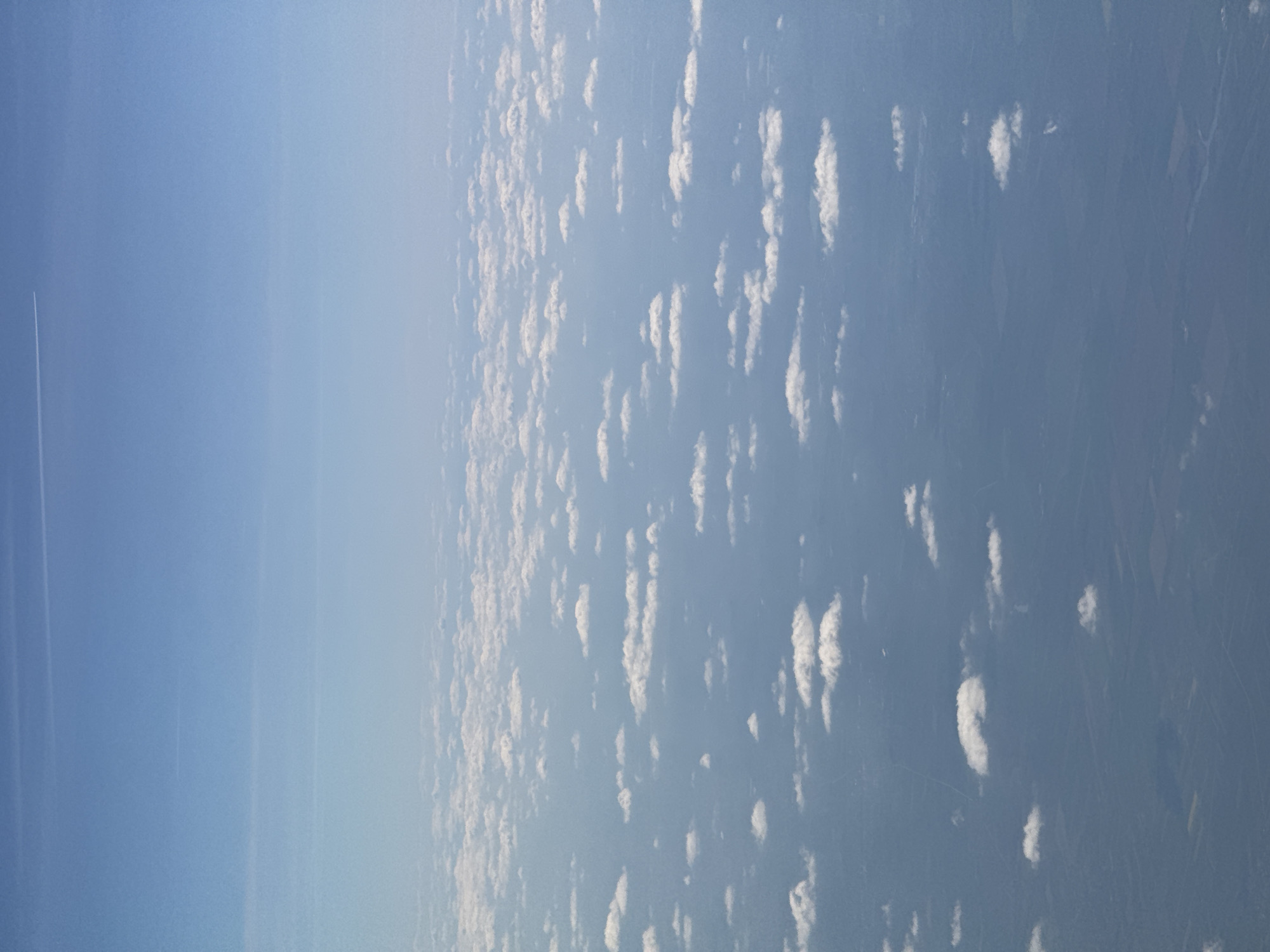 The image shows a view from an airplane over a rural area with patchy clouds. The horizon is visible on the left side. There is no discernible city in the image, so determining if it is in Sofia, Bulgaria is not possible. A contrail is visible in the upper left. The environment consists of a blue sky with sparse, elongated clouds. The land below is a mix of fields and other undefined features.