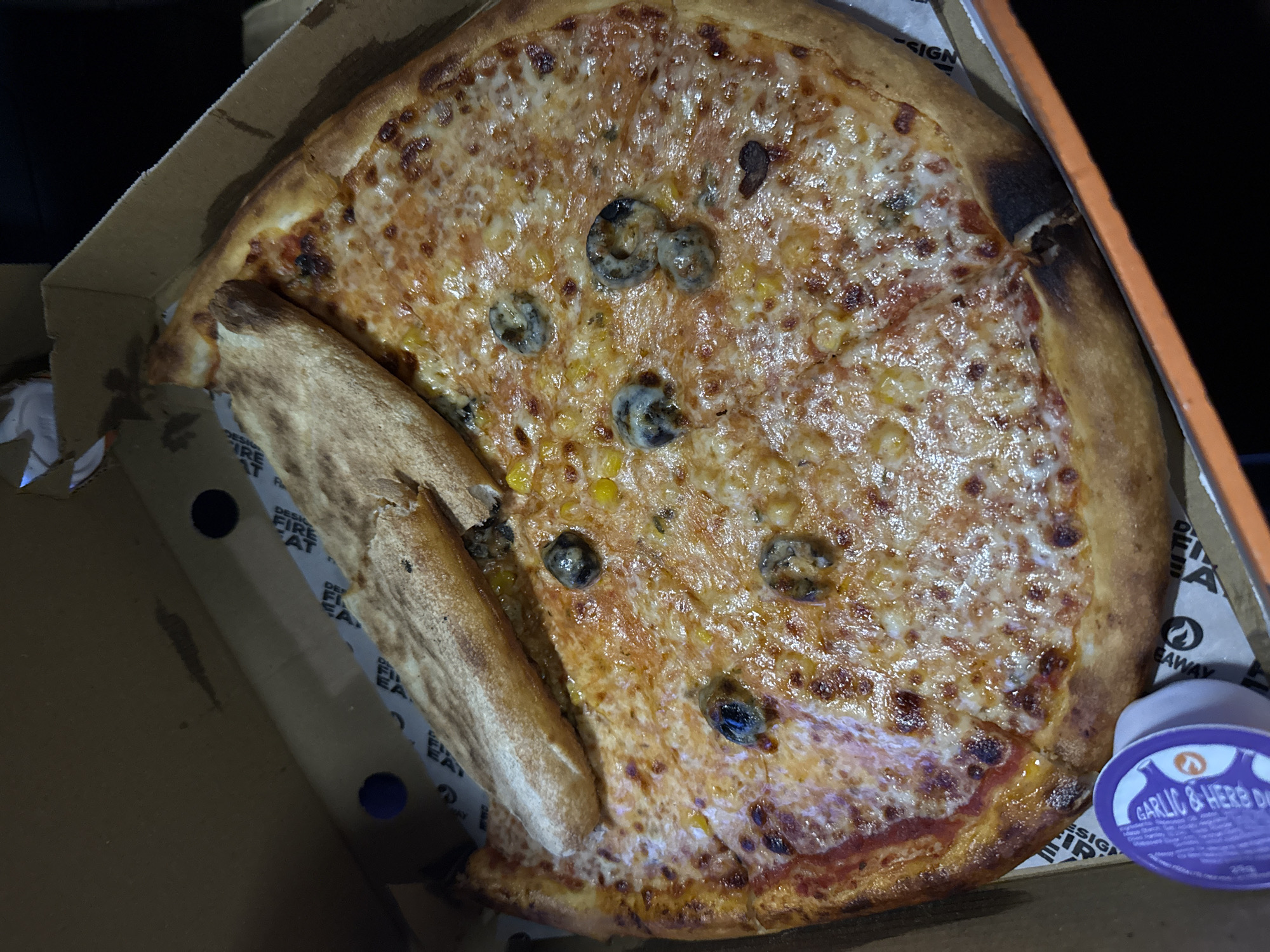 A partially eaten pizza sits in a cardboard box. A slice is detached and lies alongside the remaining pizza. The pizza is topped with cheese, corn, and what appear to be sliced black olives. A small container of garlic and herb dip is visible in the lower right corner of the box. The box indicates "Design Fire Eat" and "Takeaway" on its sides. The exact location in Rushcliffe, United Kingdom, cannot be determined from the image. No people are visible.