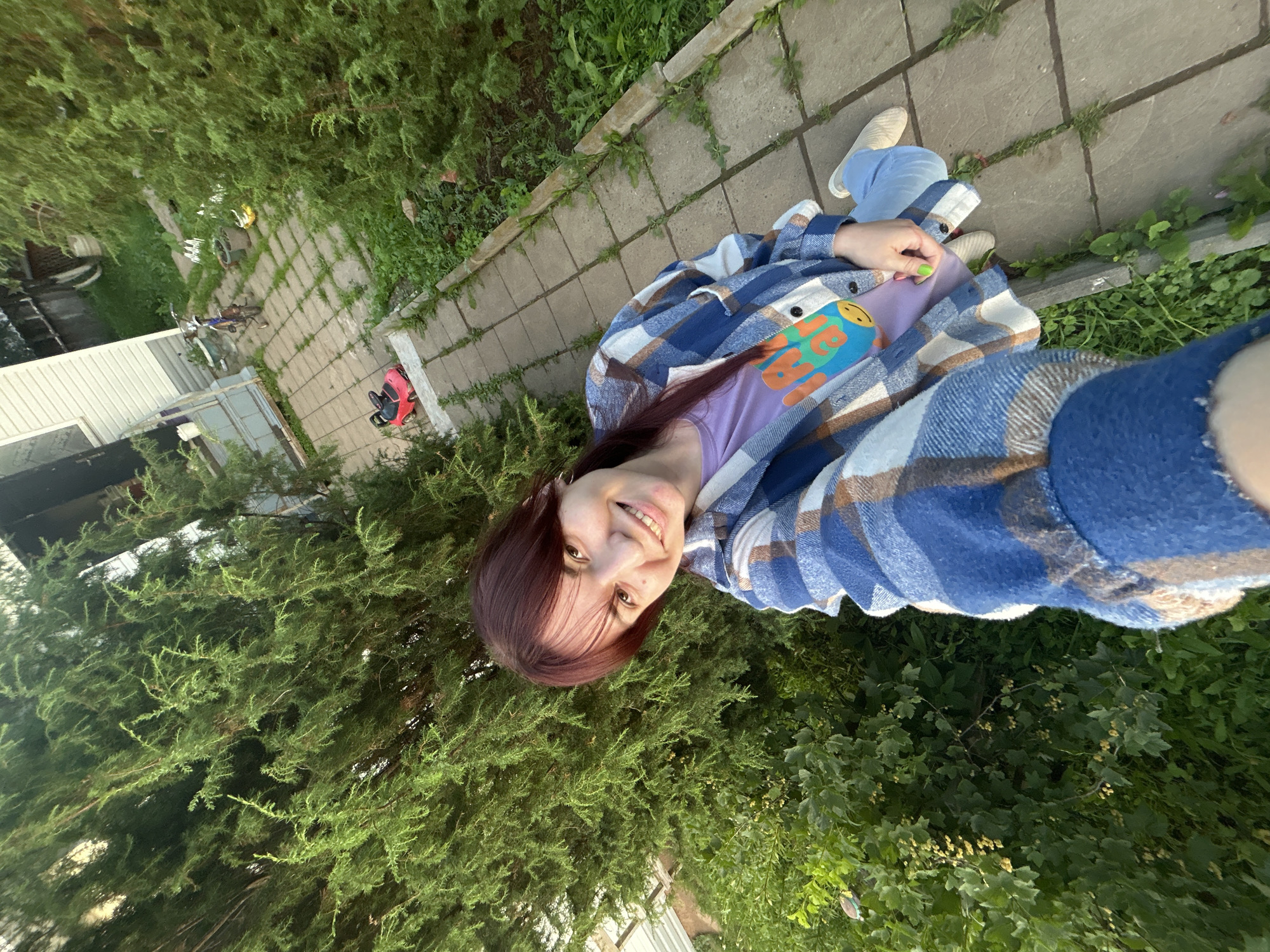 The media file shows a young woman taking a selfie outdoors. She has long, dark red hair and is wearing a purple t-shirt with a graphic print and a blue and white plaid shirt. She is smiling and looking at the camera. The setting appears to be a backyard or garden with paved walkways and lush green foliage, including trees and bushes. In the background, there's a glimpse of a white building with a corrugated metal roof, a bicycle, and a red toy vehicle. The lighting suggests it might be late afternoon or early evening, with the sun casting a warm glow. The overall mood is casual and happy. The text on the t-shirt is a graphic with cartoonish faces and lettering, but it is not legible enough to be accurately transcribed. The location is indicated as Foky, Russia.