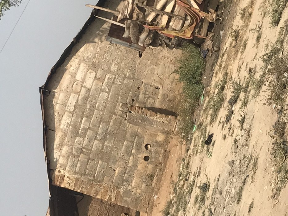 The image displays a scene in Ago-Abo, Nigeria, dominated by a single-story building constructed from unrendered concrete blocks. The visible wall exhibits significant weathering, discoloration, and minor structural wear, including a vertical section of exposed reddish brickwork and two circular holes near its base. The roof is composed of rusted, uneven corrugated metal sheets. To the right of the building, a large stack of cut timber or wooden planks is present, partially obscured by reddish-brown fabric or tarp, with a rudimentary wooden ladder leaning against it. The ground in the foreground and mid-ground is dry, light-brown earth with sparse patches of low green vegetation. On the ground, directly in front of the building, a pair of light-green flip-flops and a dark, shapeless object are visible. A light-colored chicken is partially visible on the far right, near the stacked timber. The sky overhead is a clear, light blue. No human subjects are present in the scene.