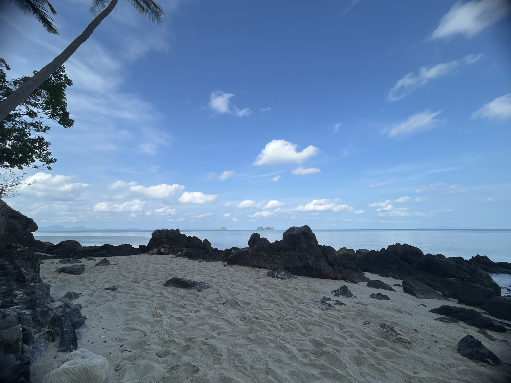 The image shows a sandy beach in Baan Taling Ngam, Thailand, with dark rocks scattered along the shore. The sky is blue with scattered white clouds. The ocean is calm and stretches to the horizon, where two small islands are visible. A palm tree is visible in the upper left corner of the image.