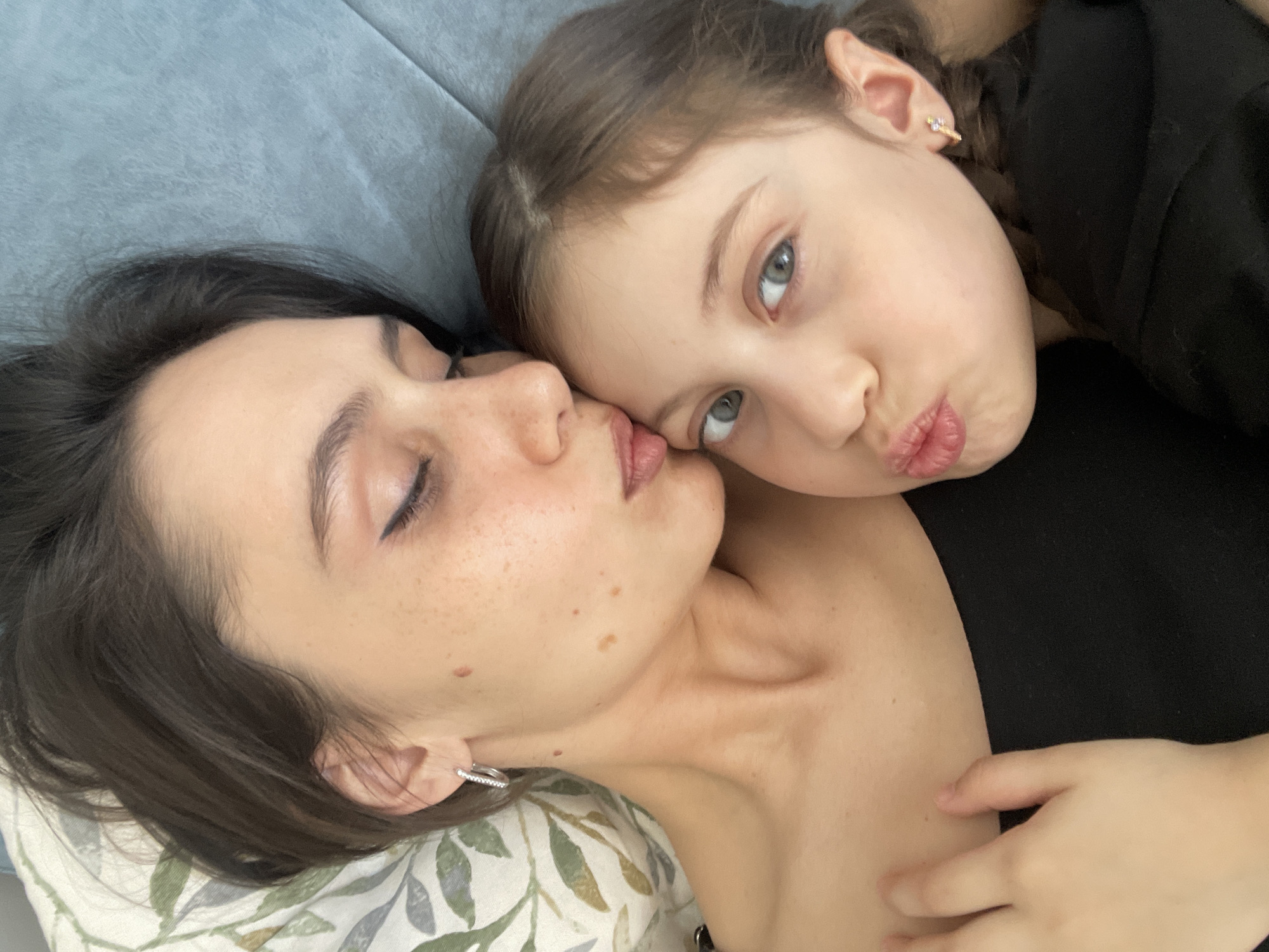 A woman and a young girl are lying down together, posing closely for a picture. The woman, who is closer to the camera, is lying on a pillow with a leaf pattern. She has dark hair and closed eyes. The girl, positioned behind the woman, is looking directly at the camera with a pouty expression. She has light brown hair and fair skin, and wears a small earring. She is wearing a black shirt. The background shows a blue couch. The photograph does not provide information about the location in the city of Izhevsk, Russia.