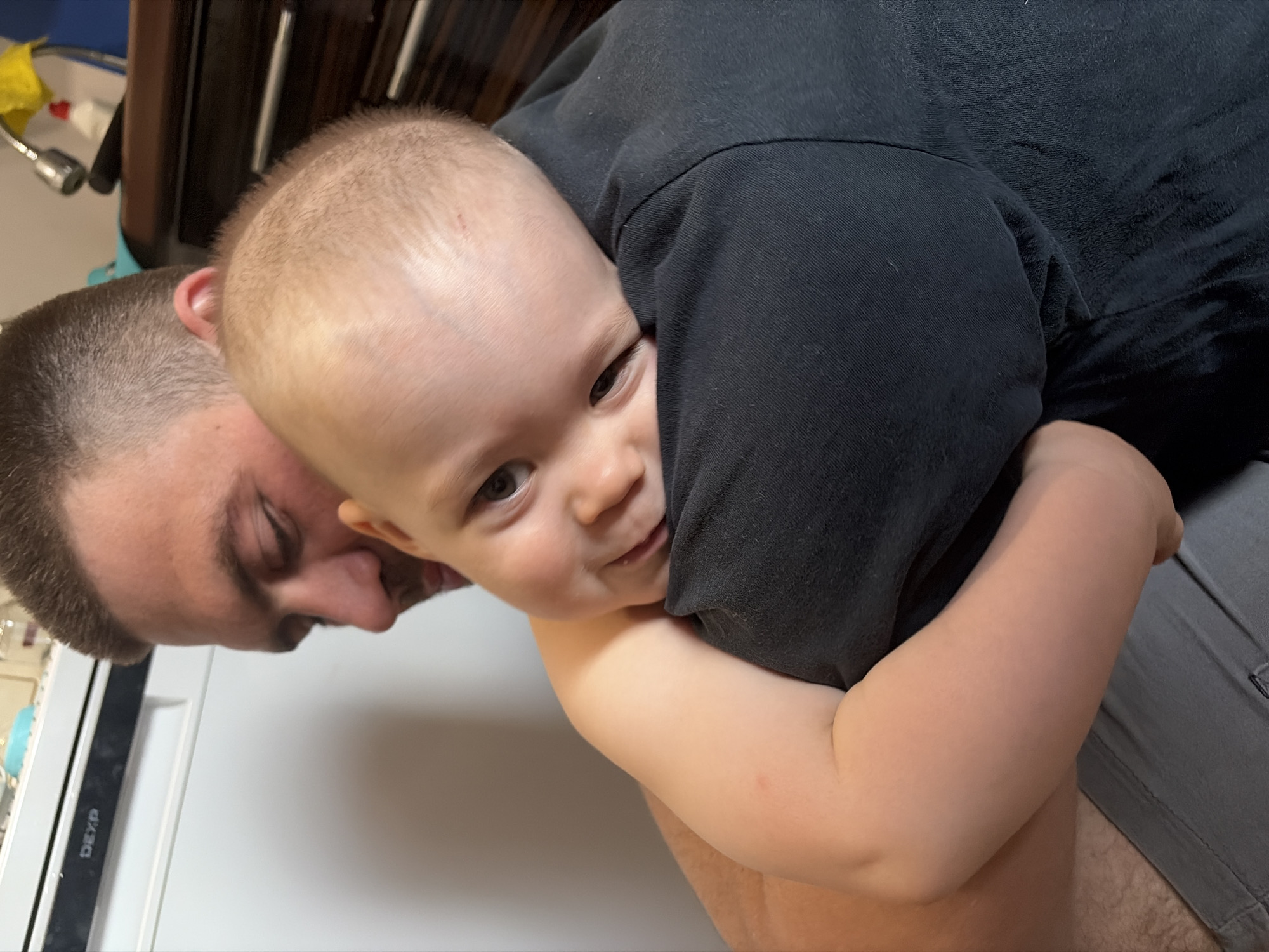 A man with closely cropped hair and a baby are embracing in an indoor setting. The man is mostly out of focus with his head tilted back. The baby's face is in focus as it nestles against the man's shoulder. The baby's arms are around the man. Part of a white refrigerator is visible in the background. It is not possible to identify if this photo was taken in Chaikovsky, Russia.