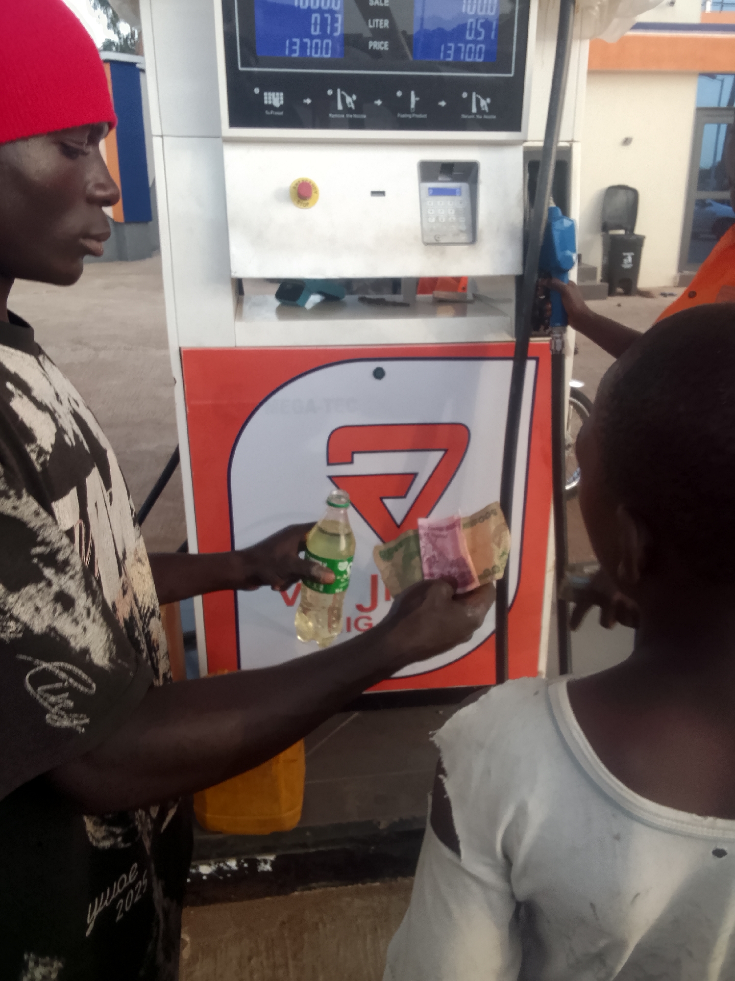 At a Vega-Tec fuel station in Kuru Dungus, Nigeria, a man wearing a red beanie and a black and white t-shirt is paying for fuel. He holds a small, clear bottle filled with a yellow liquid and several folded banknotes in his left hand. A yellow container is on the ground near his left leg. The fuel pump display reads "SALE 0.000 LITER PRICE 1370.0". Another person wearing an orange shirt is pumping fuel into a vehicle. A child is standing beside the man paying for fuel. The environment includes a paved area in front of the fuel station and a building in the background.