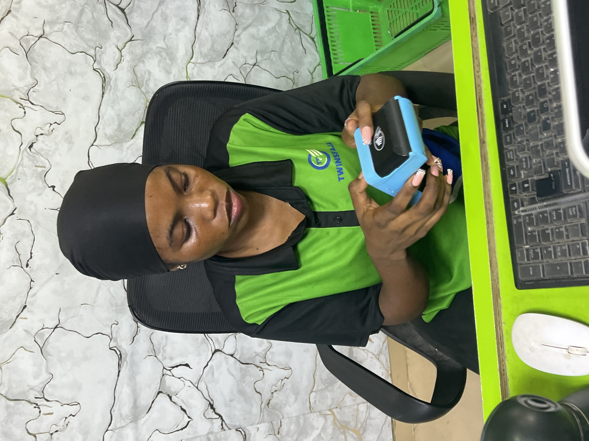 A woman in a green and black uniform with the "TWINSFAJA" logo is sitting at a desk holding a blue handheld device. The desk has a dirty keyboard, a mouse, and other items on it. There is a green basket in the background. The location is Ijegun, Nigeria.