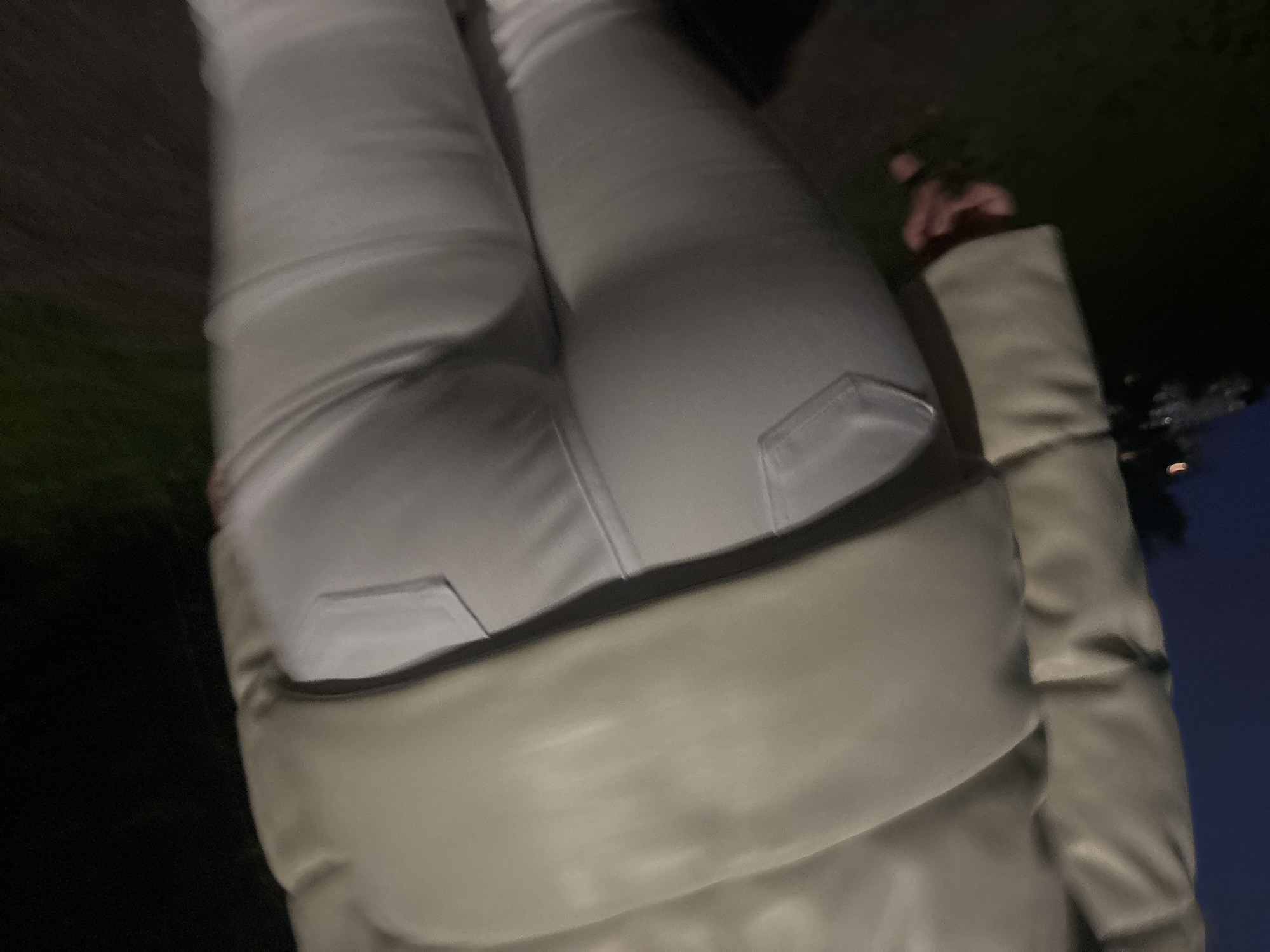 The image shows a close-up view of a person wearing white pants and a light-colored puffer jacket. The person is outdoors, likely at night, as indicated by the dark background. Some greenery is visible in the background. A portion of the person's hand is visible as well. The location is listed as Unknown, Unknown.