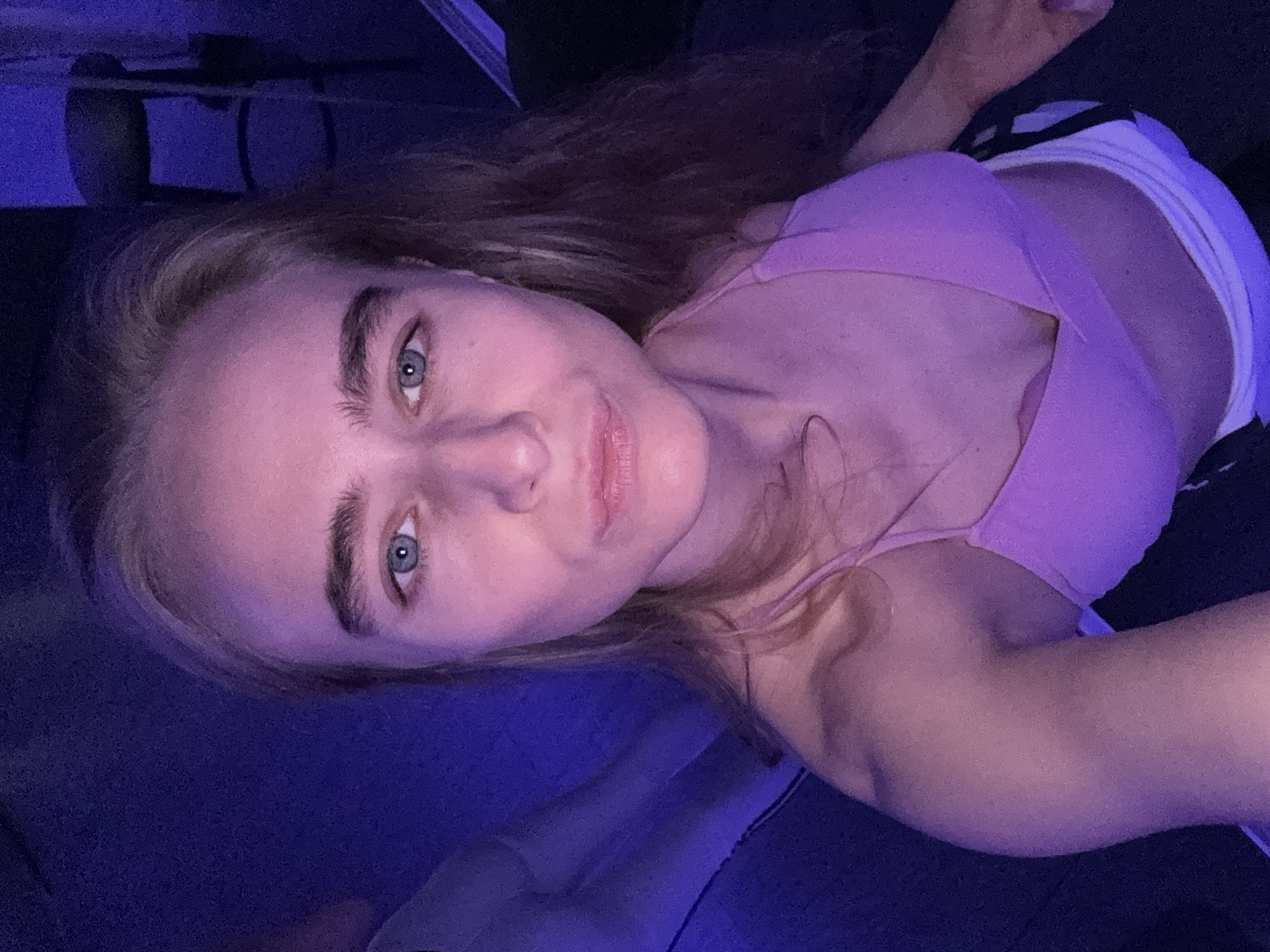 A woman with blonde hair and blue eyes is looking at the camera. She is wearing a pink top and a white garment. The scene is bathed in blue light. The location is unspecified.