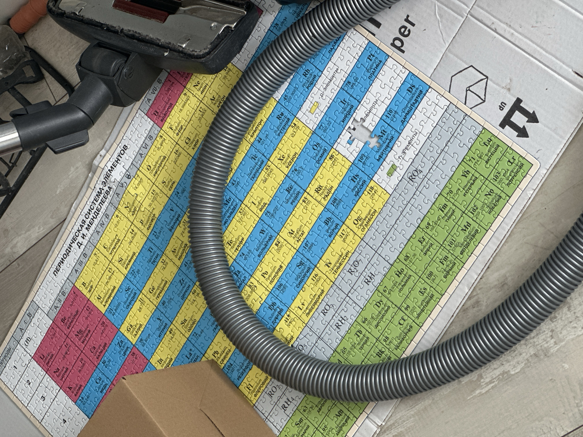 The image shows a close-up of a periodic table of elements puzzle laid out on a cardboard surface. A grey corrugated vacuum cleaner hose is coiled and positioned over a portion of the puzzle. In the background, a black vacuum cleaner attachment is visible. The scene appears to be indoors, with a wooden floor or surface visible beneath the cardboard. There is no indication of the city of Nevinnomyssk, Russia, or any people present in the image. The focus is on the juxtaposition of the scientific puzzle and the household appliance.