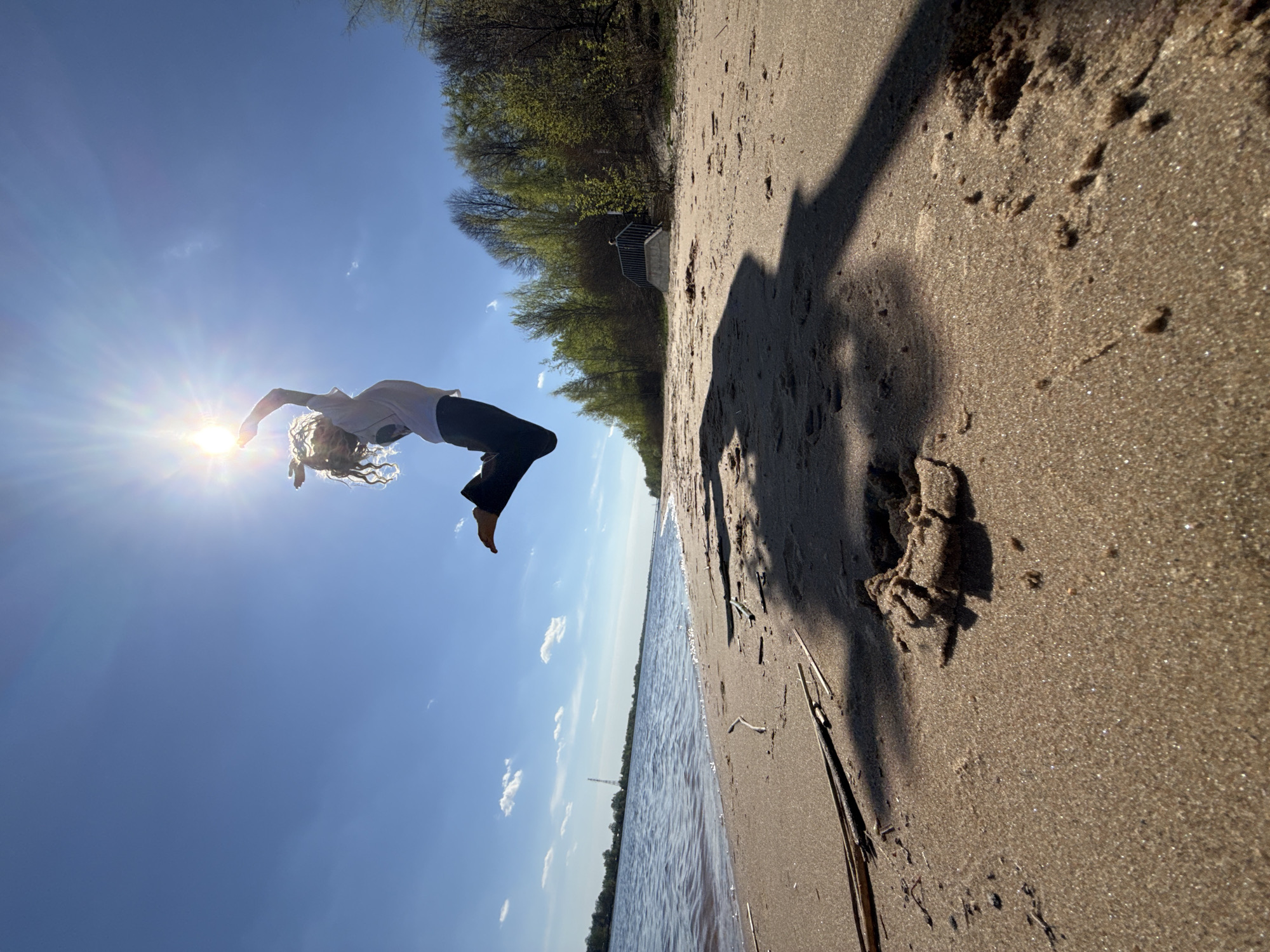 A person is performing a backbend on a beach near a body of water on a sunny day. The person is silhouetted against the bright sun in the upper left of the frame, wearing a light-colored shirt and dark pants, with blond hair. The beach consists of sand, with shadows cast upon it. Vegetation and a stairway can be seen along the edge of the beach. The water stretches towards the horizon under a blue sky with scattered clouds.
Based on the prompt's location request, this image is likely taken on a beach in Rusanovo, Russia, though this cannot be confirmed with certainty from visual information alone.