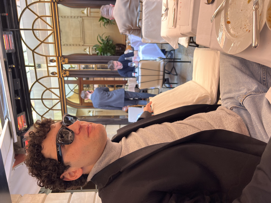 The image depicts an upscale dining scene at Café Costes, part of Hôtel Costes, located at 239 Rue Saint Honoré, Paris, France. The primary subject is a young adult male, positioned in the foreground and facing slightly to his left. He has dark, curly hair and wears black Ray-Ban Wayfarer sunglasses, a light grey turtleneck sweater, a dark blazer, and light blue jeans. His right hand rests on the back of a nearby empty chair with a white cushion. On the white-linened table in front of him, a white plate containing residual food and cutlery (fork and knife) is visible. A menu displaying "COSTE" is also on the table.
In the midground and background, other patrons are present in the semi-enclosed restaurant space. To the right, a man with light hair is seated facing away from the camera, wearing a light brown suit jacket. Further back and slightly to the left, another man in a dark long-sleeved top is seated, looking down at his table which holds a wine glass and a bottle. To the far left in the background, a man in a grey suit stands, seemingly using a mobile phone. The architectural setting features light-colored stone walls, ornate gold-colored column details, and large arched glass panels or windows, suggesting a grand, classic Parisian interior courtyard or terrace. Overhead, several red infrared heating lamps are mounted. Green potted plants enhance the sophisticated ambiance. The floor is a light-colored tile.