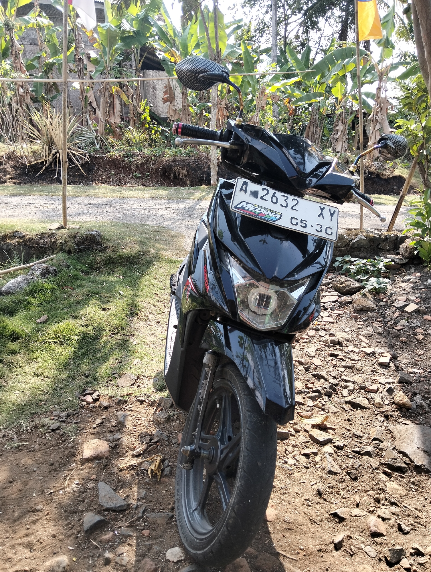 The image shows a black motorcycle parked on a dirt and rocky ground, with a grassy area to the left. The motorcycle is facing forward, with its front wheel and headlight clearly visible. The license plate reads "A 2632 XY G5 3C". In the background, there are banana trees, other foliage, and a wooden fence. There are also two flags visible, one red and white and another yellow. The scene appears to be in a rural or semi-rural area, possibly in Karangnunggal, Indonesia, given the presence of banana trees and the style of the license plate. The lighting suggests it is daytime, with sunlight casting shadows. No people are visible in the image.
