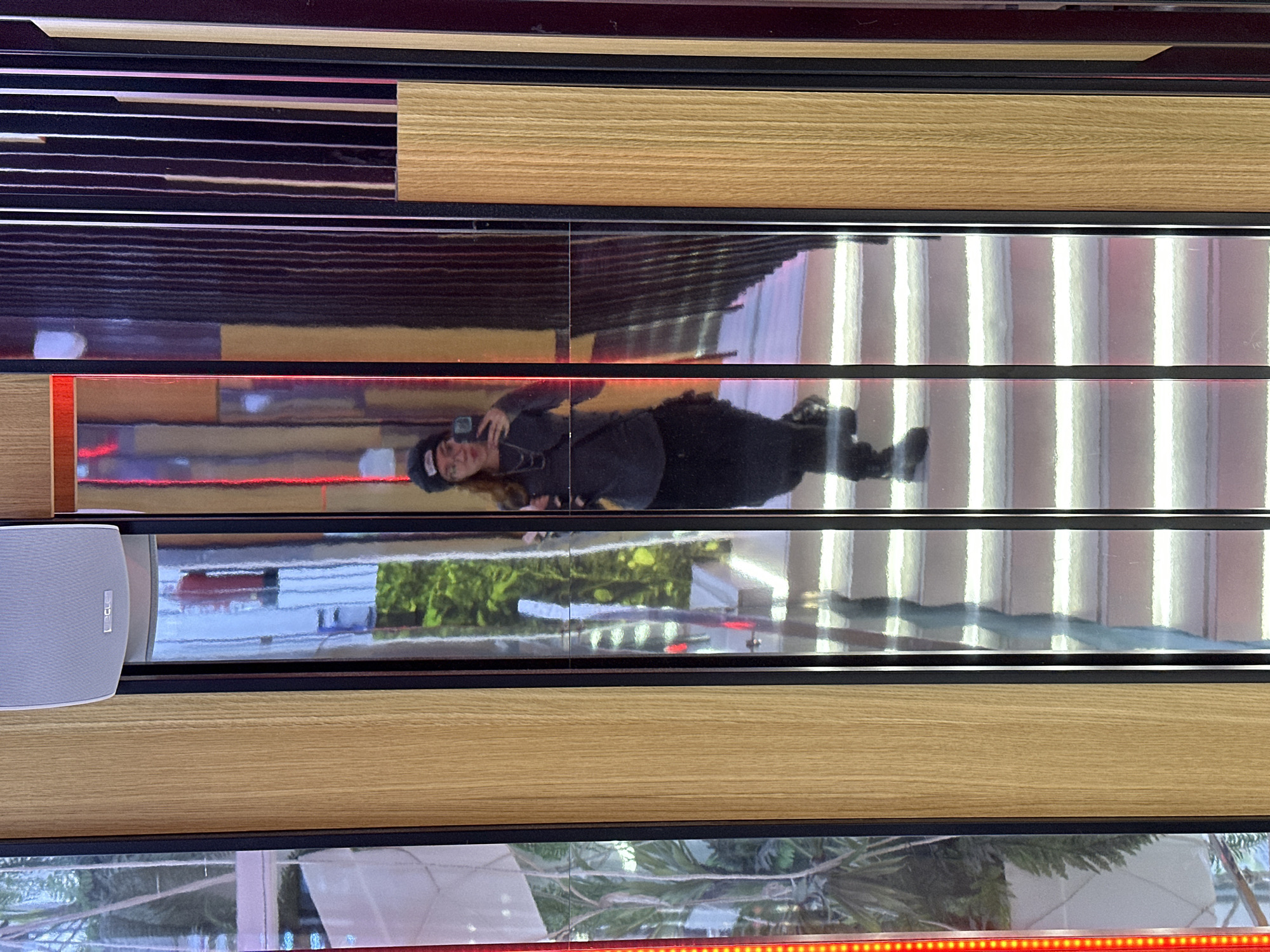 The image displays a series of horizontal strips, alternating between highly reflective surfaces, natural light-colored wood, and dark, solid bands. The image itself appears to be rotated 90 degrees clockwise, influencing the perceived orientation of the elements.
In one of the central, mirror-like strips, a person is prominently reflected, appearing to lie horizontally across the frame. They are dressed in dark clothing, including a long-sleeved top, pants, and a cap or beanie. The individual is holding a smartphone, seemingly capturing a photograph or video of their reflection or the surrounding environment. Their face is partially visible.
Other reflective strips show distorted elements of the scene, including bright vertical light sources and patches of green foliage, suggesting trees or plants, indicating either an outdoor view or an indoor space with significant natural light. A light grey speaker, bearing the text "Lectron," is mounted on one of the wooden strips on the left side of the image. Additionally, strips of red LED lighting are visible, bordering some of the reflective sections, particularly at the bottom of the frame and near the person's reflection.
The composition of reflective surfaces, wood, and lighting suggests a modern interior or an art installation, possibly within a building in Barcelona, Spain. The diverse materials and the play of light create a dynamic and abstract visual experience.