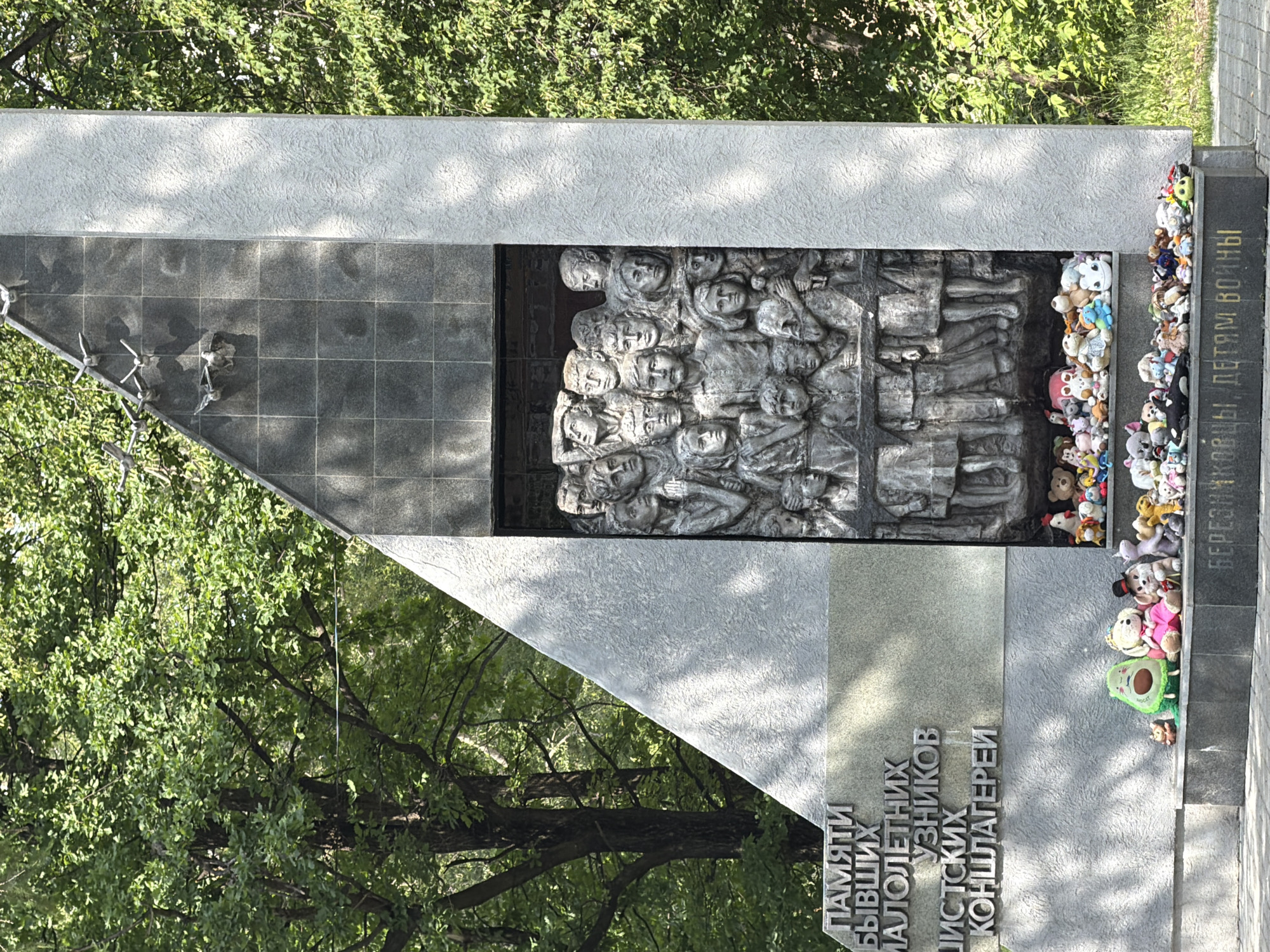 A monument stands outdoors, surrounded by lush green trees. The monument has a gray stone base, with a large triangular section to the left and a tall rectangular section to the right. The triangular section features inscribed text in Russian. The rectangular section has a dark gray stone inset with a bas-relief sculpture of a crowd of children and adults, their faces and bodies etched in stone, appearing to look upwards. Above the bas-relief, a series of metal bird sculptures are attached to the dark gray stone, seeming to fly away. At the base of the monument, a collection of colorful stuffed animals, including bears, rabbits, and other plush toys, are arranged on the steps and ledges. A horizontal inscription in Russian, "БЕРЕЗНИКОВЦЫ, ДЕТЯМ ВОЙНЫ" (Berezniki residents, to the children of war), is visible on the front of the base. The scene is illuminated by natural sunlight, casting shadows on the monument and the surrounding ground. The image is captured from a slightly upward angle, emphasizing the scale of the monument against the backdrop of the trees.
