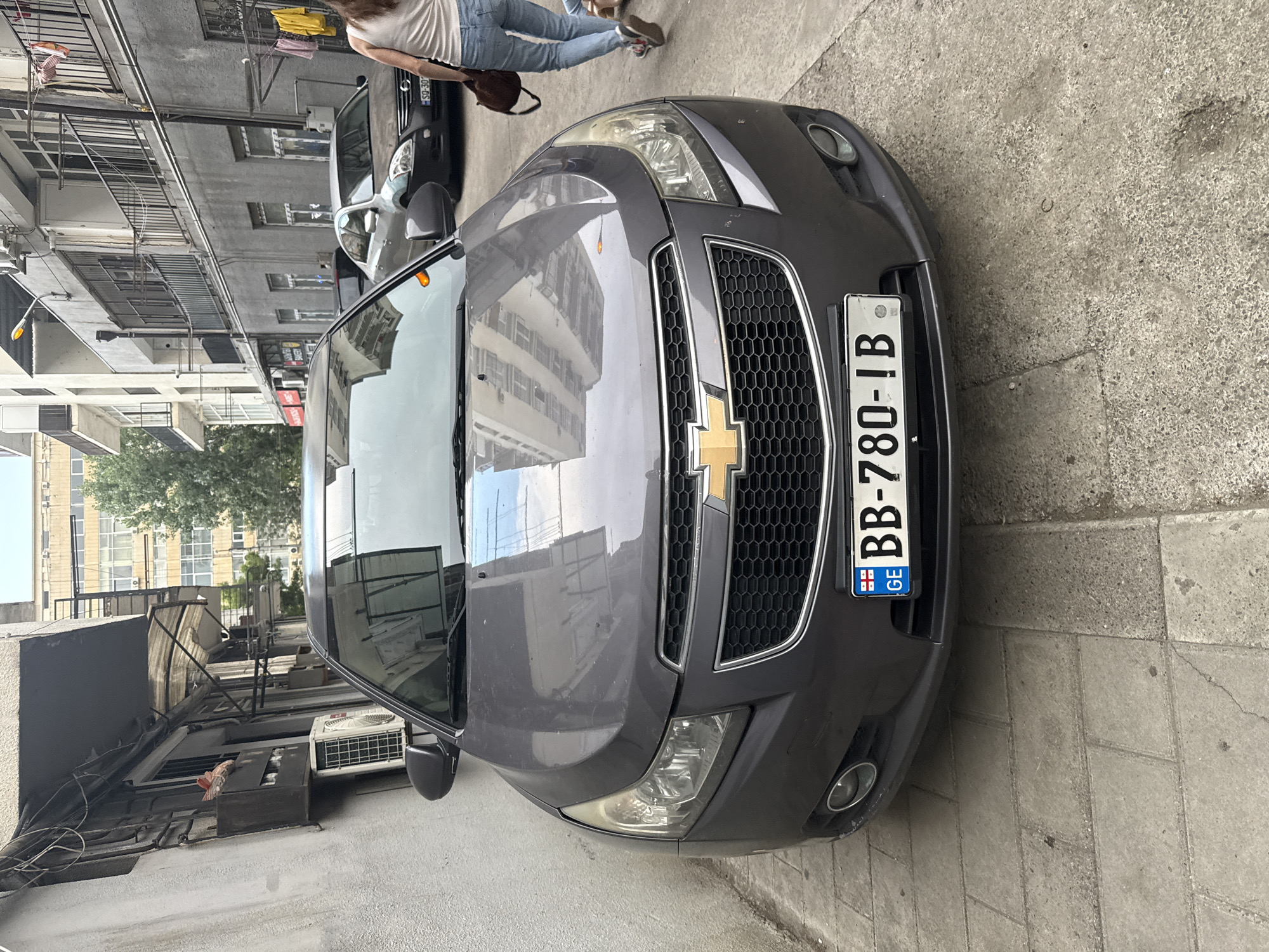 This image shows the front of a dark gray Chevrolet parked on a paved street in Tbilisi, Georgia. The license plate reads "BB-780-IB" with a Georgian flag emblem. To the left, a white car is parked behind the Chevrolet. In the background, a person is walking away from the camera, wearing a white top and blue jeans. The buildings on either side of the street appear to be apartment blocks, some with balconies and air conditioning units visible. The time of day appears to be daytime, with natural light illuminating the scene. The weather seems clear. The overall scene depicts a typical urban street environment in Tbilisi.