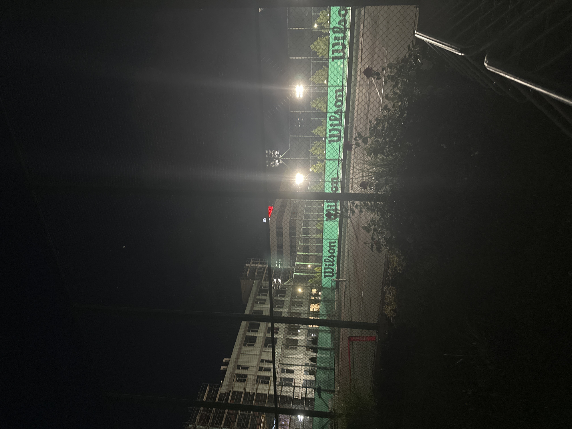 A nighttime shot of an outdoor tennis court complex in Sofia, Bulgaria. The court is illuminated by floodlights. A chain-link fence encloses the court. Beyond the fence, a multi-story building, possibly under construction due to scaffolding, is visible. The building appears to be lit from within. The tennis court fencing has "Wilson" branding printed on it. In the foreground, dark vegetation and a section of a metal railing are visible. The sky is dark, suggesting it is late at night.