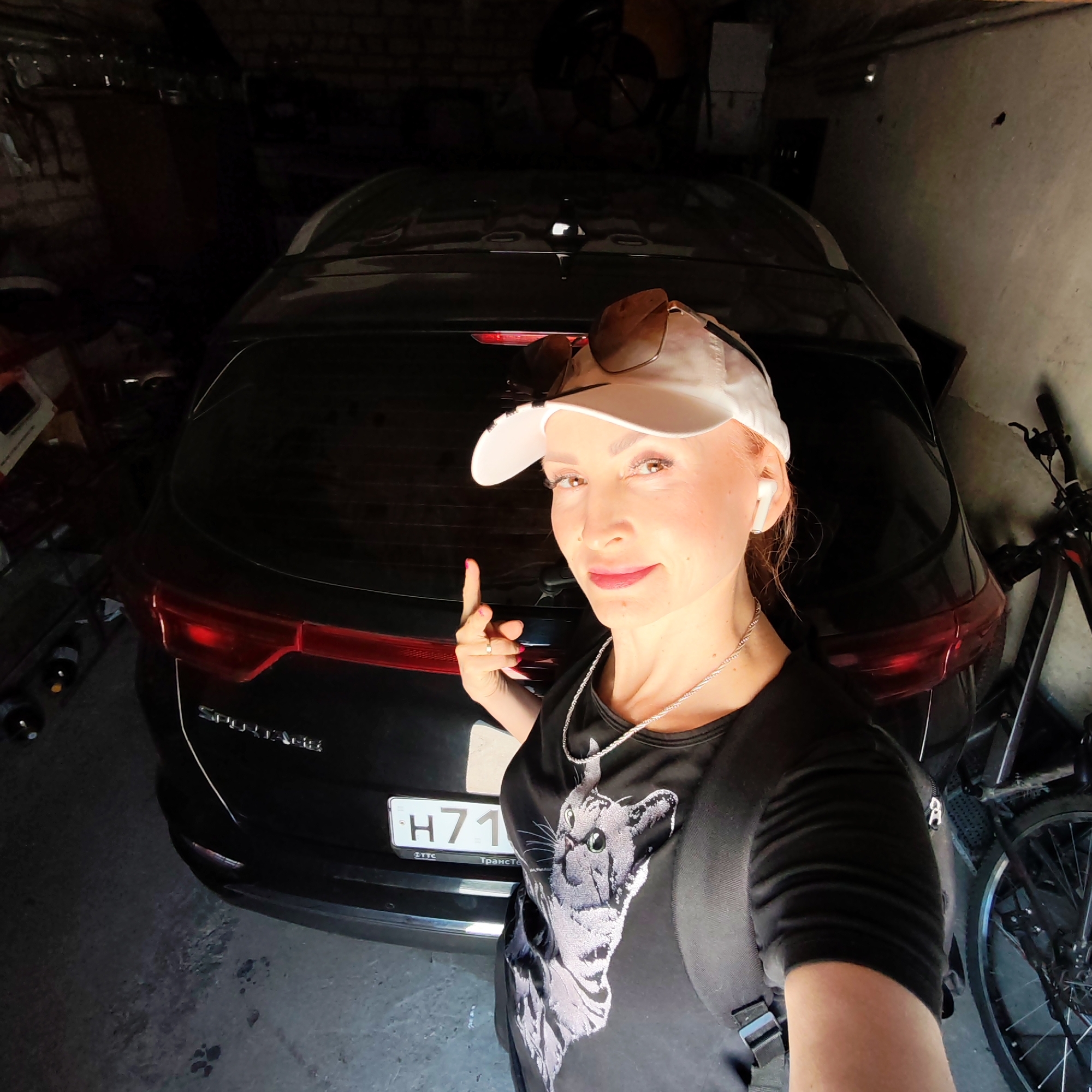 A selfie taken by a woman in a garage. The woman is Caucasian, with fair skin, red hair, and a white baseball cap with sunglasses perched on top. She's wearing a black t-shirt with a cat design, a silver necklace, and a black backpack. An earbud is visible in her right ear. She is pointing with her right index finger towards the back of a black SUV, a Kia Sportage, parked in the garage. The license plate of the car is visible and appears to read "H71", potentially a Russian license plate. To the right of the car, a bicycle is visible. The garage has a concrete floor with what appear to be paw prints. The back wall of the garage is made of brick. Miscellaneous items are visible in the background. The garage appears to be located in Chaikovsky, Russia, based on the car's registration details.