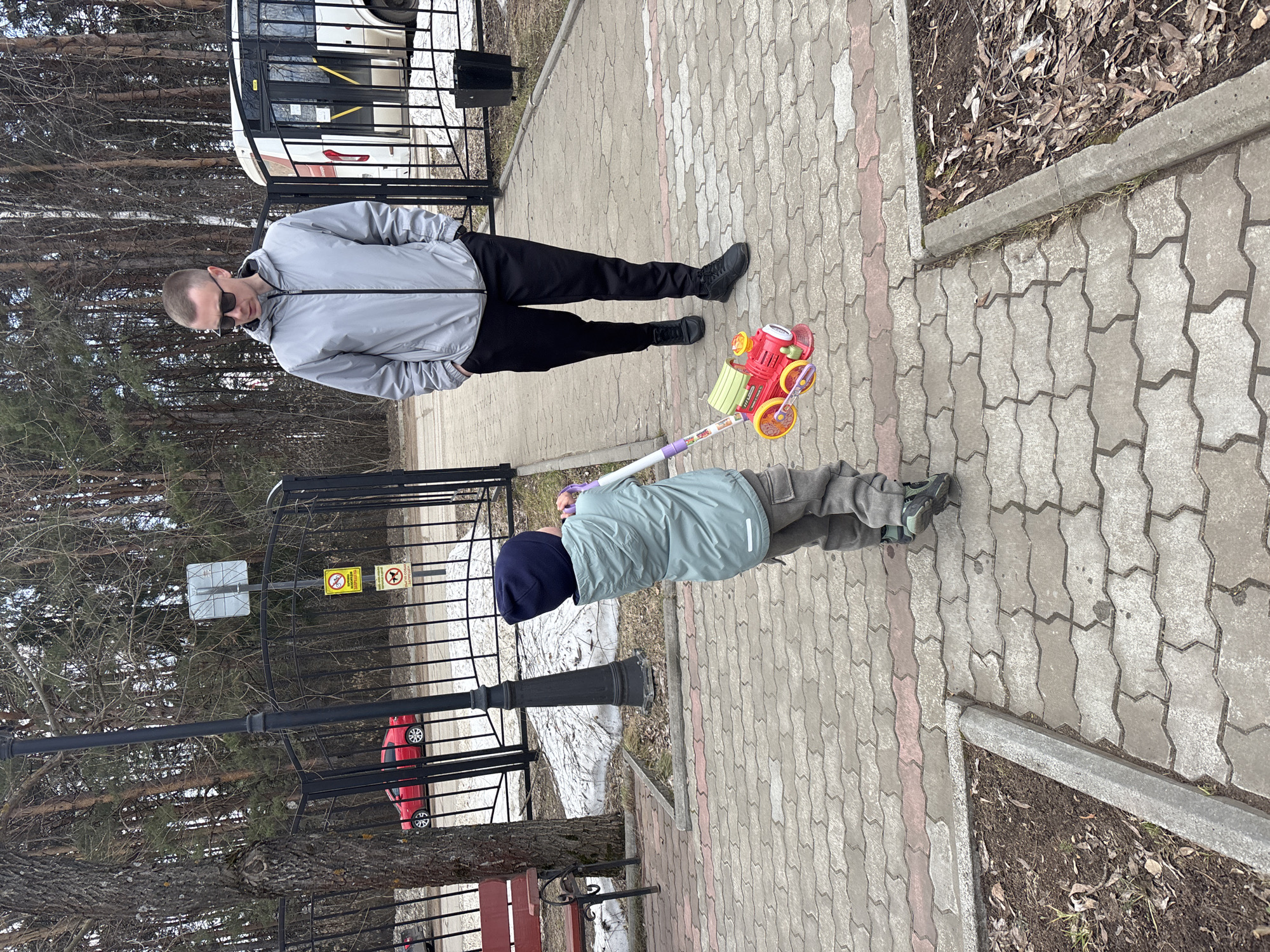 This image, taken in Chaikovsky, Russia, depicts an outdoor scene featuring an adult male and a young child on a paved walkway. The photo is rotated 90 degrees counter-clockwise from its original orientation.
The adult male stands in the foreground, facing slightly to the right of the frame. He wears a light grey hooded jacket, black pants, and black shoes, and has short hair and sunglasses. His hands appear to be in his pockets. The young child is further down the path, facing away from the camera. The child wears a light blue-green jacket, grey pants, and a dark blue beanie, and is pushing a brightly colored red and yellow wheeled toy with a long handle.
The walkway is made of interlocking concrete pavers, predominantly grey with some reddish-brown sections. To the left of the walkway is a wooded area with numerous bare trees, suggesting late winter or early spring. To the right, a strip of ground with dried leaves and some grass is visible, bordered by a concrete curb.
In the background, behind a black metal fence, a white bus with yellow stripes and a red car are partially visible. Patches of snow can be seen on the ground beyond the fence. A black, ornamental lamppost stands near the fence. Two yellow warning signs are attached to the fence: one with a crossed-out dog symbol (indicating "no dogs") and another with a crossed-out bicycle or scooter symbol (indicating "no bicycles/scooters").
The scene takes place during the daytime, under soft, diffused light, consistent with cool weather. The overall impression is a casual outdoor outing, with the child playing with a toy.