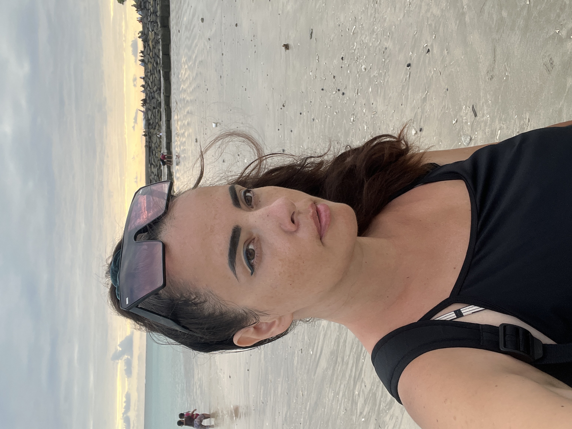 A woman takes a selfie on a beach in Kuta, Indonesia. She is in the foreground, facing slightly to the right, wearing a black tank top and a backpack. She has dark hair pulled back and sunglasses perched on her head. In the background, the ocean meets the shore, with a few people visible in the water. Further back, a rocky pier extends into the water, with a line of people and a small plane visible in the distance. The sky is overcast with hints of yellow and orange from a sunset.