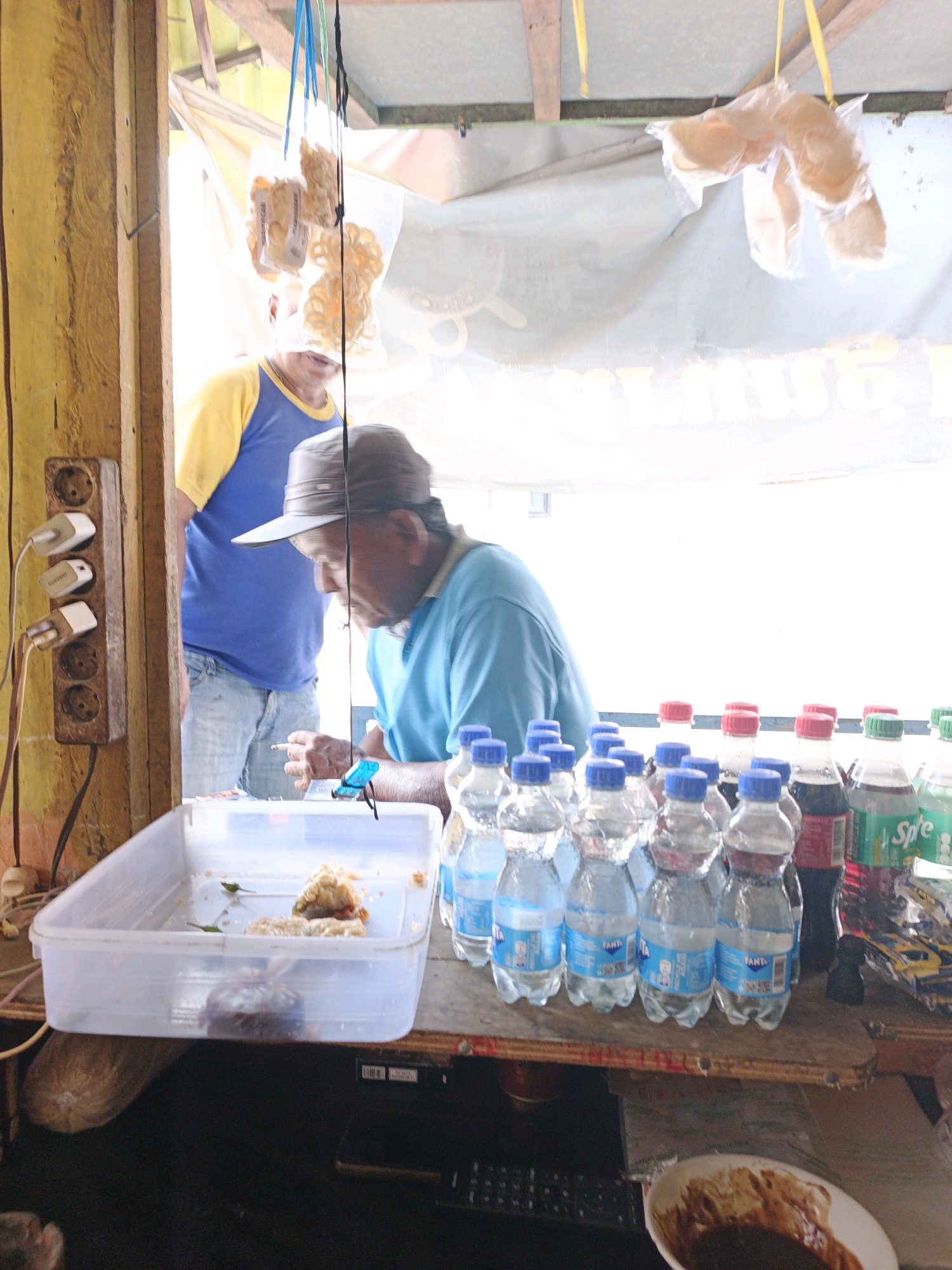The image captures a street-side stall, likely in Ketegan, Indonesia, with a focus on the transactions and offerings. A man wearing a blue shirt and a hat is leaning over a counter, holding a cigarette and appearing to be interacting with a customer or examining goods. To his left, another individual in a blue and yellow shirt stands partially visible, suggesting a shared space or adjacent stall. The counter displays an assortment of beverages, predominantly bottled water and soda, with a clear plastic container holding what appears to be fried snacks. In the background, translucent plastic bags containing more snacks hang from the ceiling. An electrical outlet strip with multiple plugs is affixed to a wooden post on the left side of the frame. The overall scene suggests a bustling commercial environment.