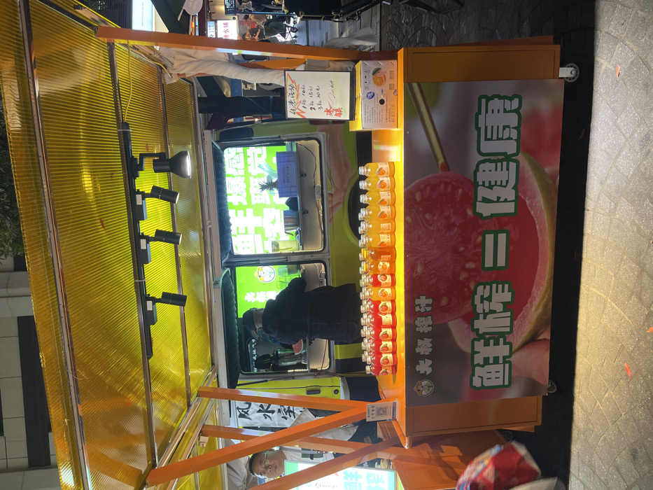 A yellow food cart with a yellow awning sits on a sidewalk in Hang Zhou Shi, China (latitude: 30.264, longitude: 120.156). The cart is displaying rows of small glass bottles containing a bright red liquid, possibly guava juice. The bottles are arranged in front of a large, colorful advertisement featuring a guava fruit with the Chinese characters "Guava = Health" on a yellow background. A man wearing a black jacket is standing beside the cart. The cart is parked in front of a larger, white food truck with a yellow sign that reads "Guava" and depicts a picture of a guava fruit. The truck has a window and interior that is partially visible and appears to be stocked with various items. The scene is illuminated by streetlights, casting a yellow glow on the sidewalk.