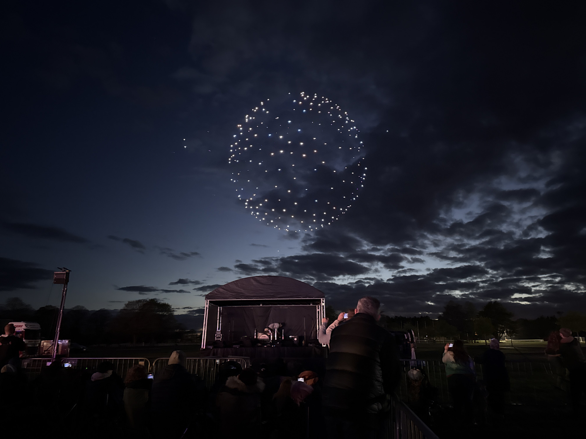 The image depicts a dark outdoor event, likely a concert or performance, in what is possibly South Norfolk, United Kingdom. A stage with a black canopy is visible in the middle ground, containing a drum set and other equipment. A crowd of people stands in the foreground, with many holding up phones to record the event. Above the stage, in the night sky, is a formation of lights arranged in an oval or circular shape. The sky is filled with dark, textured clouds. The overall lighting is dim, creating a nighttime atmosphere. The presence of a truck and other equipment suggests that this event is taking place in a park or open space. A metal barrier separates the crowd from the stage.