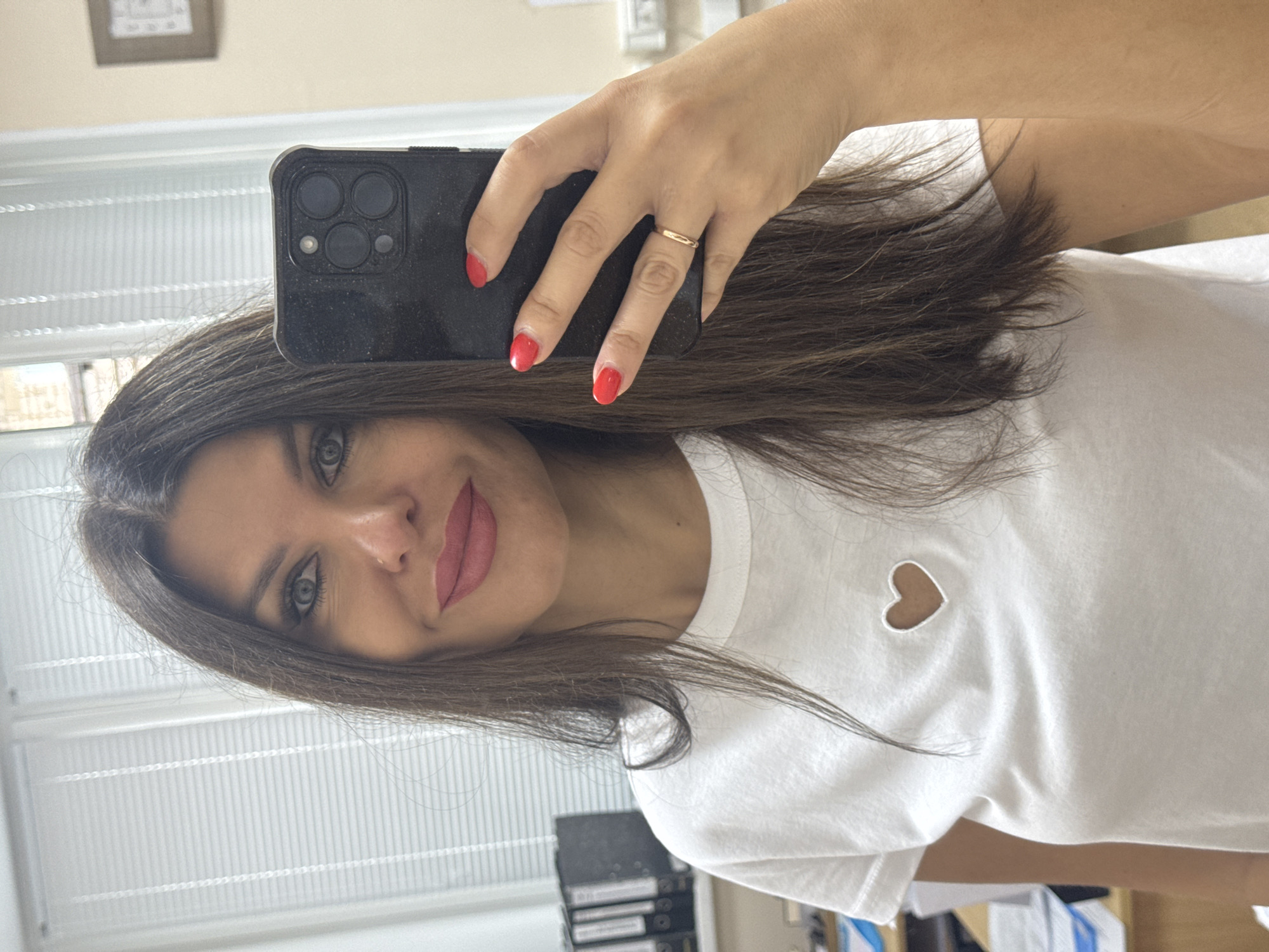 A woman is taking a selfie in an office. She has long, dark brown hair, blue eyes, and is wearing pink lipstick. She is wearing a white t-shirt with a heart cut-out on the chest. Her left hand is holding a black smartphone, and she is wearing a gold ring on her ring finger and red nail polish. The background appears to be an office with blinds on a window, filing cabinets, and a framed picture on the wall. The image does not provide any specific information to determine its location in Chaikovsky, Russia.