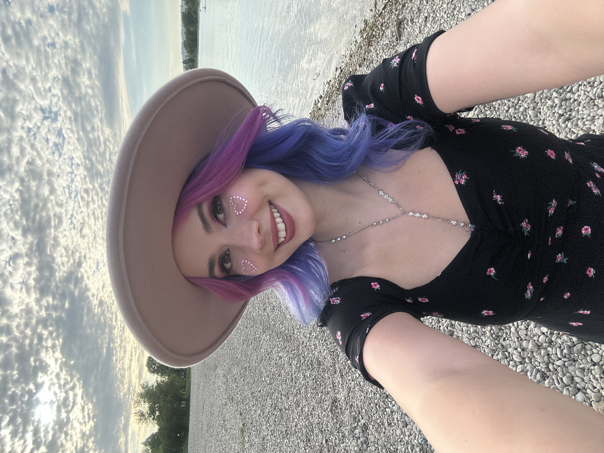 A young woman is featured in a vibrant selfie, taken outdoors. She smiles broadly at the camera, her head tilted slightly to her left. Her distinctive hair transitions from a bright pink at the roots to a deep blue-purple towards the ends. She wears a wide-brimmed, light brown fedora-style hat. Her cheeks are adorned with sparkling, heart-shaped glitter stickers, and she has visible freckles. She is dressed in a black short-sleeved dress with a pattern of small pink flowers, and a silver-toned necklace with small, round beads or charms. Her right arm is visible in the foreground, likely holding the camera for the selfie.
The background depicts a tranquil outdoor setting, consistent with a lakeside or riverside location, such as Jarun Lake in Zagreb, Croatia. The immediate foreground and mid-ground consist of a pebbly or gravelly shore. Beyond this, a calm body of water extends towards the horizon, reflecting the soft light. The sky above is partly cloudy, displaying scattered white clouds against a pale blue and subtly golden hue, indicating either late afternoon or early evening. The woman's joyful expression and the serene environment evoke a sense of happiness and leisure. There is no visible text in the image.