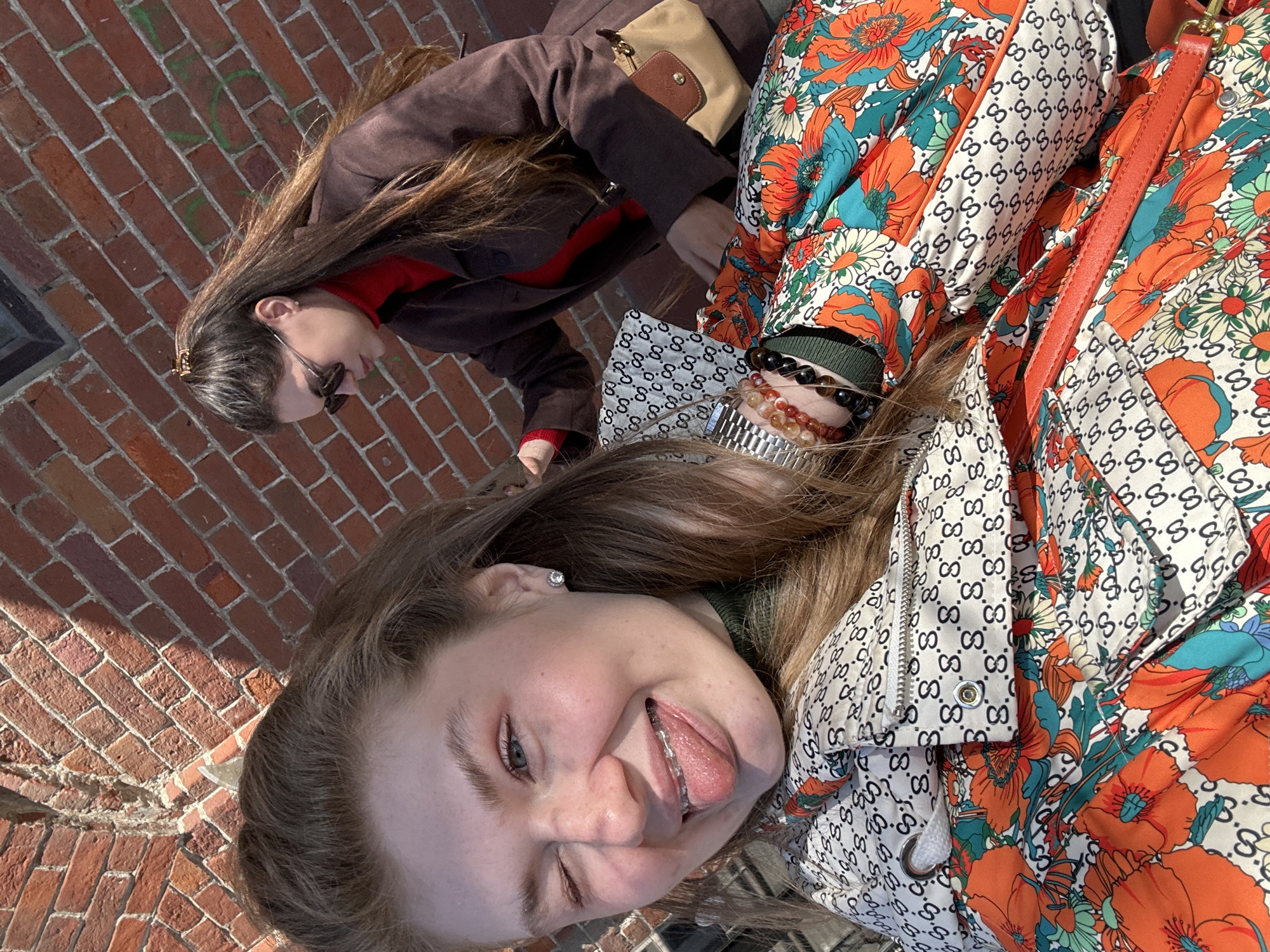 Two young women are posing outdoors. The woman in the foreground is sticking her tongue out and winking. She is wearing a printed jacket. The second woman, wearing sunglasses and a brown jacket, is behind the first. The background features a brick wall. This scene takes place in Kazan, Russia.