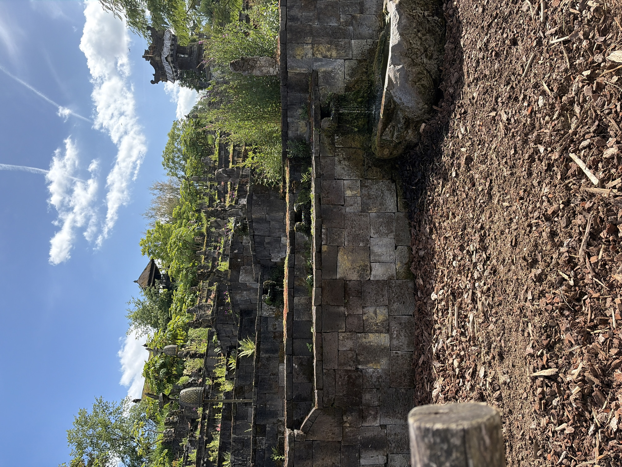 The image depicts the Balinese garden at Pairi Daiza park in Cambron-Casteau, Belgium. It is a sunny day with a bright blue sky and scattered clouds. The foreground is filled with dark brown wood chips covering the ground. A thick, weathered wooden log is positioned in the lower left corner, partially obscuring the view of the scene beyond.
The midground features a multi-tiered stone structure that resembles a terraced hillside. The stone blocks are of various sizes and appear weathered, with some covered in moss and algae. Lush green vegetation, including ferns and flowering plants, grows in the crevices and on the terraces of the stone structure. Scattered among the greenery are numerous small stone statues, some appearing to be of human or animal figures. In the mid-right, a small waterfall cascades over moss-covered stones into a stone basin, part of which is a large, rough-hewn boulder.
The background shows more of the terraced garden, with taller trees and a traditional Balinese structure with a thatched roof visible at the top of the terraces. Another ornate, temple-like structure with intricate carvings is also present on the right side of the background. The overall impression is one of a serene, exotic, and meticulously landscaped garden.