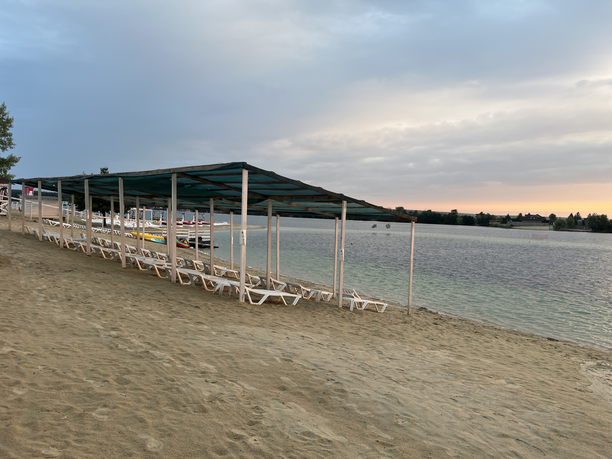 The image depicts a sandy beach area next to a body of water, likely a lake or reservoir, in Bezliudivka, Ukraine. Several rows of empty white lounge chairs are neatly arranged on the sand under a long, covered structure with a green tarp roof supported by wooden poles. Beyond the chairs and the covered area, the tranquil water stretches towards a distant treeline. The sky above is overcast with hints of soft, warm light suggesting either sunrise or sunset. The environment appears peaceful and prepared for visitors, though no people are present in the frame.