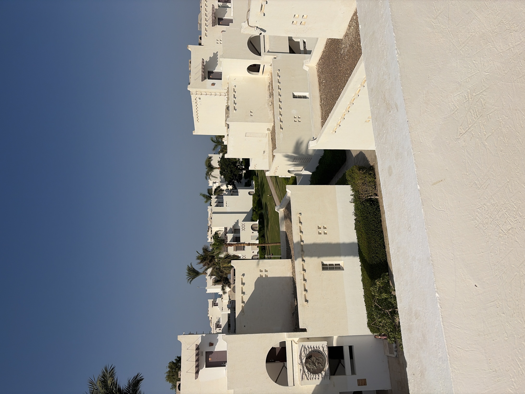 The image captures a section of a resort complex, likely in Hurghada, Egypt, characterized by its distinct architectural style. The buildings are predominantly white, featuring flat roofs, angular designs, and arched doorways and windows. Many structures have multiple levels and are interconnected or clustered together. Small, square windows are visible on some of the building facades, along with decorative horizontal elements. A few palm trees are scattered throughout the complex, adding a touch of tropical greenery. The ground surrounding the buildings appears to be a mix of green lawns, paved walkways, and areas covered with gravel or small stones, especially on the rooftops. The sky is a clear, deep blue, indicating a sunny day. In the foreground, a white, textured wall with a rough surface occupies the lower portion of the image, suggesting a vantage point from another building or elevated area. The overall impression is one of a well-maintained, sunny resort environment. No people are visible in the image.