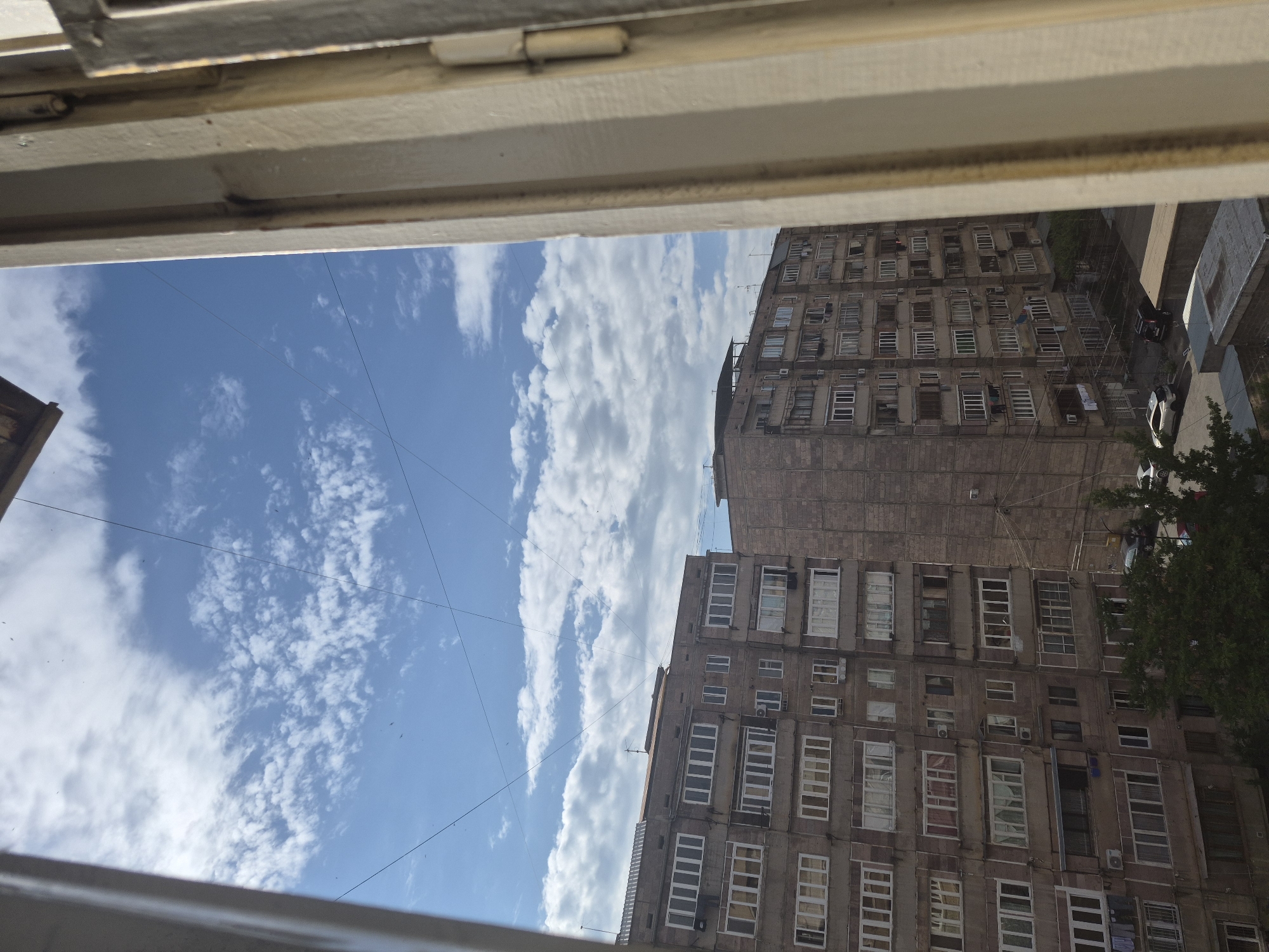This media file captures an urban residential scene, likely viewed from a window, as indicated by the prominent beige window frames visible along the top and bottom edges of the image. The perspective is rotated 90 degrees clockwise.
**Subjects:**
The primary subjects are two multi-story apartment buildings and a bright blue sky filled with scattered white cumulus clouds. The building on the upper right is a tall, greyish structure, likely made of concrete, featuring numerous windows and balconies. Many windows have white frames, some with external security bars or closed blinds. Several air conditioning units are mounted on its facade. A second, shorter apartment building is partially visible on the lower right. On the ground level between the buildings, a dark green tree with lush foliage is visible in the lower right corner, and several cars, including a white sedan and a black vehicle, are parked or driving on what appears to be a street or parking area. Thin, dark utility wires crisscross the sky on the left side of the frame. No people are explicitly visible.
**Activity or event taking place:**
The image depicts a static moment of everyday urban life. No specific activity or event is actively occurring.
**Notable details:**
The scene is set during the daytime, evidenced by the bright sunlight illuminating the clouds and buildings. The weather appears clear and fair, with a vivid blue sky and scattered, fluffy white clouds. The architectural style of the buildings, with their dense arrangement of windows and balconies, is characteristic of urban residential areas, consistent with locations like Yerevan, Armenia. The buildings show some signs of age or wear. There is no visible text in the image. The overall impression is one of a typical, peaceful urban environment.