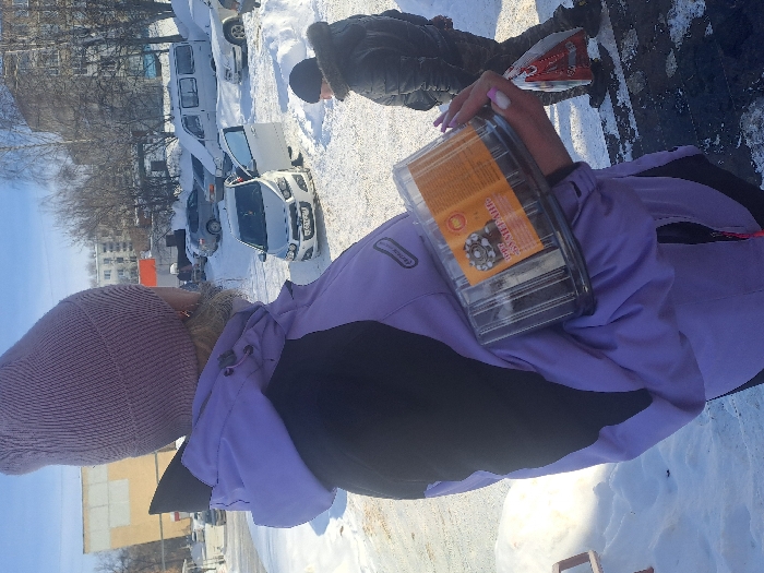 The image displays a clear winter day scene in Bolshaya Elhovka, Russia. In the foreground, a person is viewed from behind and slightly to the right, wearing a light purple knit beanie and a purple and black winter jacket with a visible fur collar. This individual holds a clear plastic container, bearing a label with Cyrillic text including "ТОРТ" (CAKE), in their right arm; their fingernails are painted a light pink. The ground is heavily covered in snow, with some areas of visible asphalt. In the mid-ground, a white Chevrolet Aveo sedan is parked on the snow, its driver's side door open. The vehicle's Russian license plate contains the characters "K 471 CX 71". To the right of the Chevrolet, another person, seen from behind, walks rightward, wearing a dark winter jacket with a fur-lined hood and carrying a red and white plastic bag in their left hand. The background features several other parked vehicles, including a white van partially covered in snow, and multi-story buildings, one with a light yellow and white facade, under a clear blue sky. Bare deciduous trees are visible in the distance.