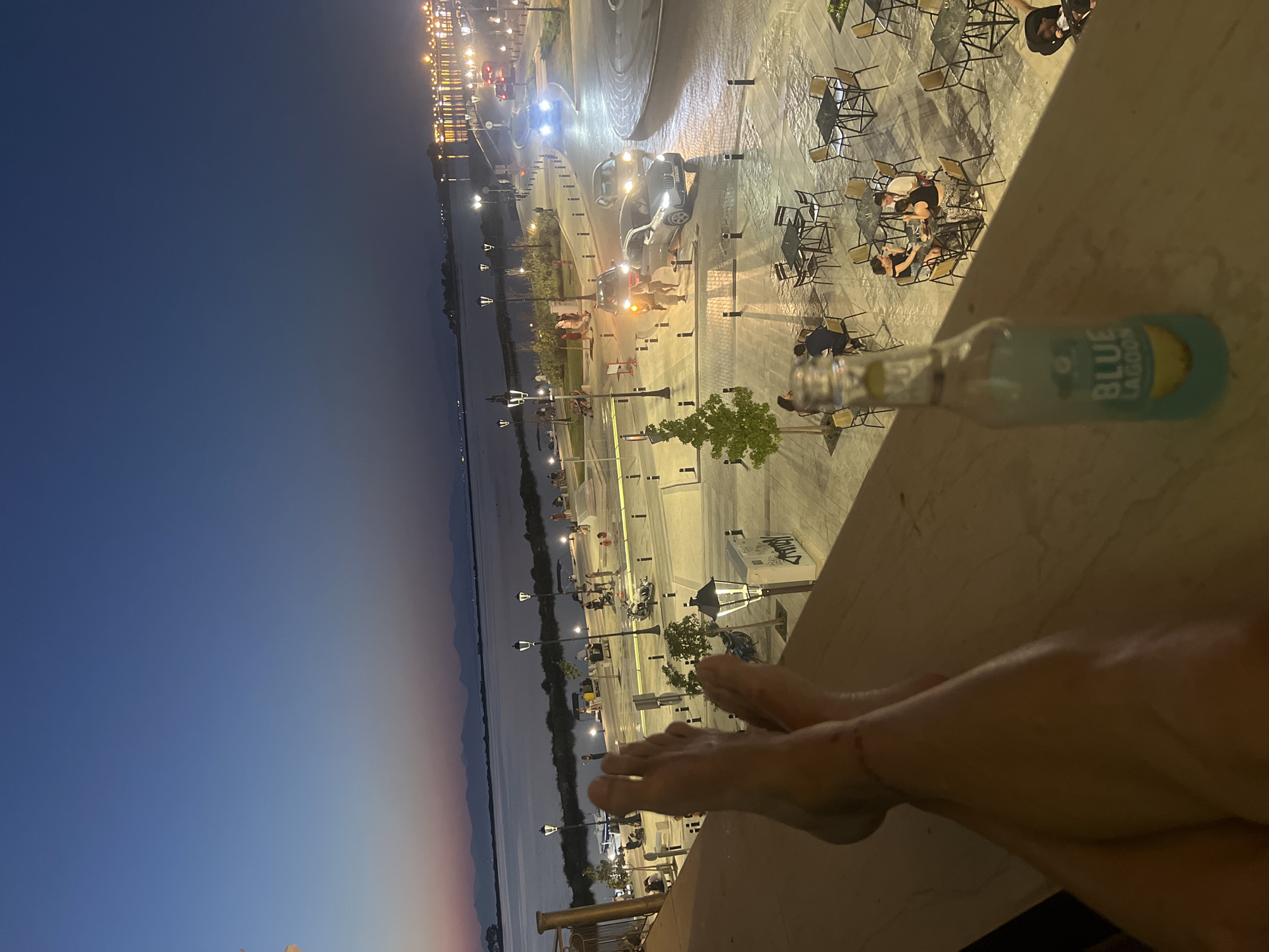 In this medium shot, taken at dusk from an elevated vantage point, a person's legs and feet are prominently positioned in the foreground, resting on a ledge. The legs are bare, and a delicate anklet is visible on the left ankle. To the right of the legs, a clear glass bottle with a blue liquid and a label reading "BLUE LAGOON" is placed on the ledge.
The background reveals a vibrant city scene. A body of water, likely the Ionian Sea given the location, stretches across the mid-ground, with distant mountains silhouetted against the twilight sky. The shoreline is lined with lampposts and vegetation, and a promenade with people strolling is visible. Beyond the promenade, a paved area features numerous outdoor dining tables and chairs, some occupied by patrons. Cars are driving on a street that curves through the scene, illuminated by streetlights and headlights. The architecture in the far background suggests a coastal town with illuminated buildings. The overall impression is of a lively evening in a seaside municipality, possibly within the Municipal Unit of Lefkada, Greece, where people are enjoying the transition from day to night.