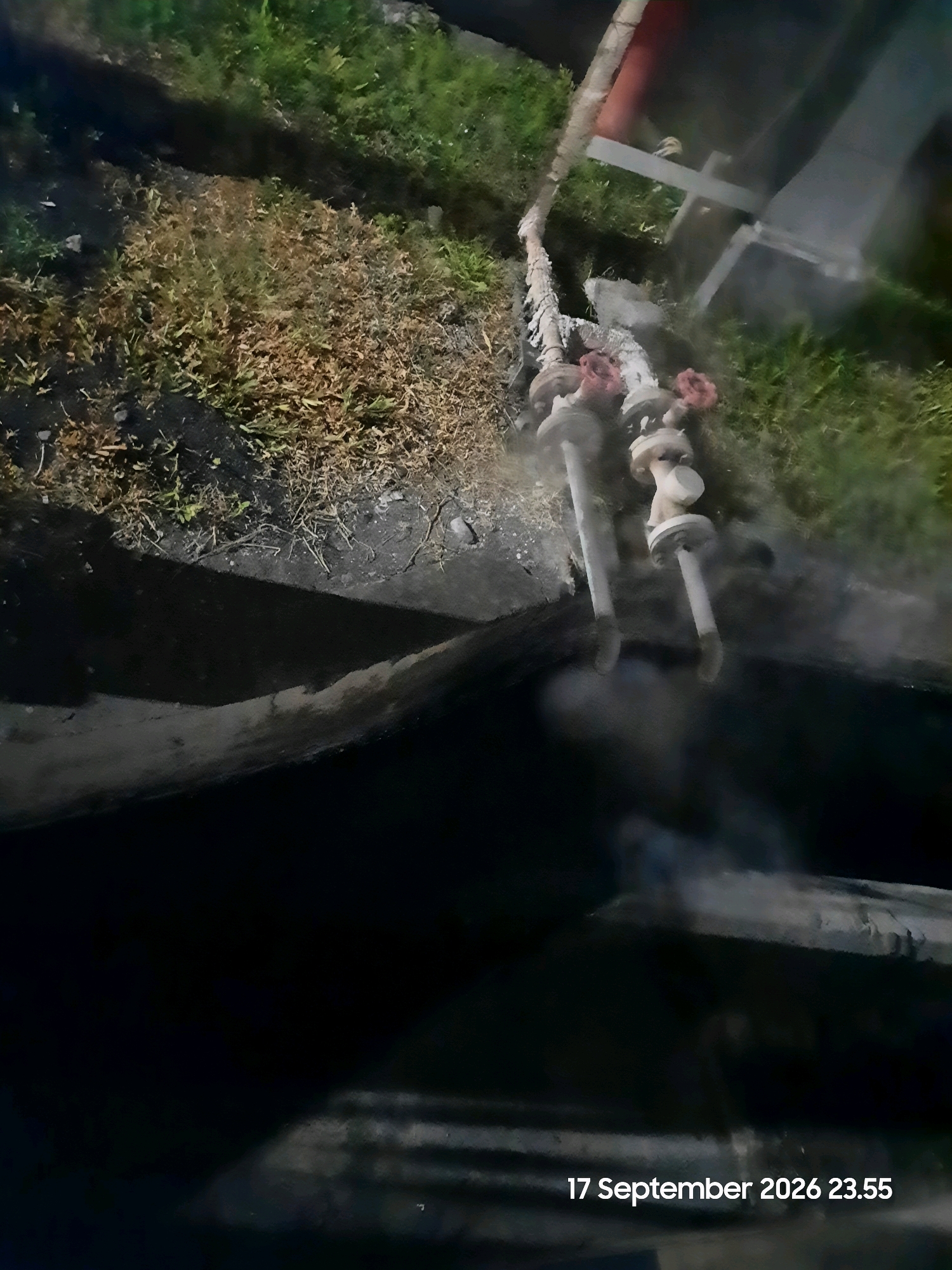 The image shows a close-up view of a fire hose connected to two spigots, with smoke rising from the hose and the ground. The hose appears to be made of rough, woven material and is lying across a patch of dry grass and soil. The spigots are metallic and have red handles. In the background, there is some blurred greenery and a structure that could be part of a building or equipment. The bottom right corner of the image displays a timestamp: "17 September 2026 23.55". The overall scene suggests an active fire-fighting or emergency situation, likely at night, given the lighting and timestamp. The location is identified as Gending, Indonesia.