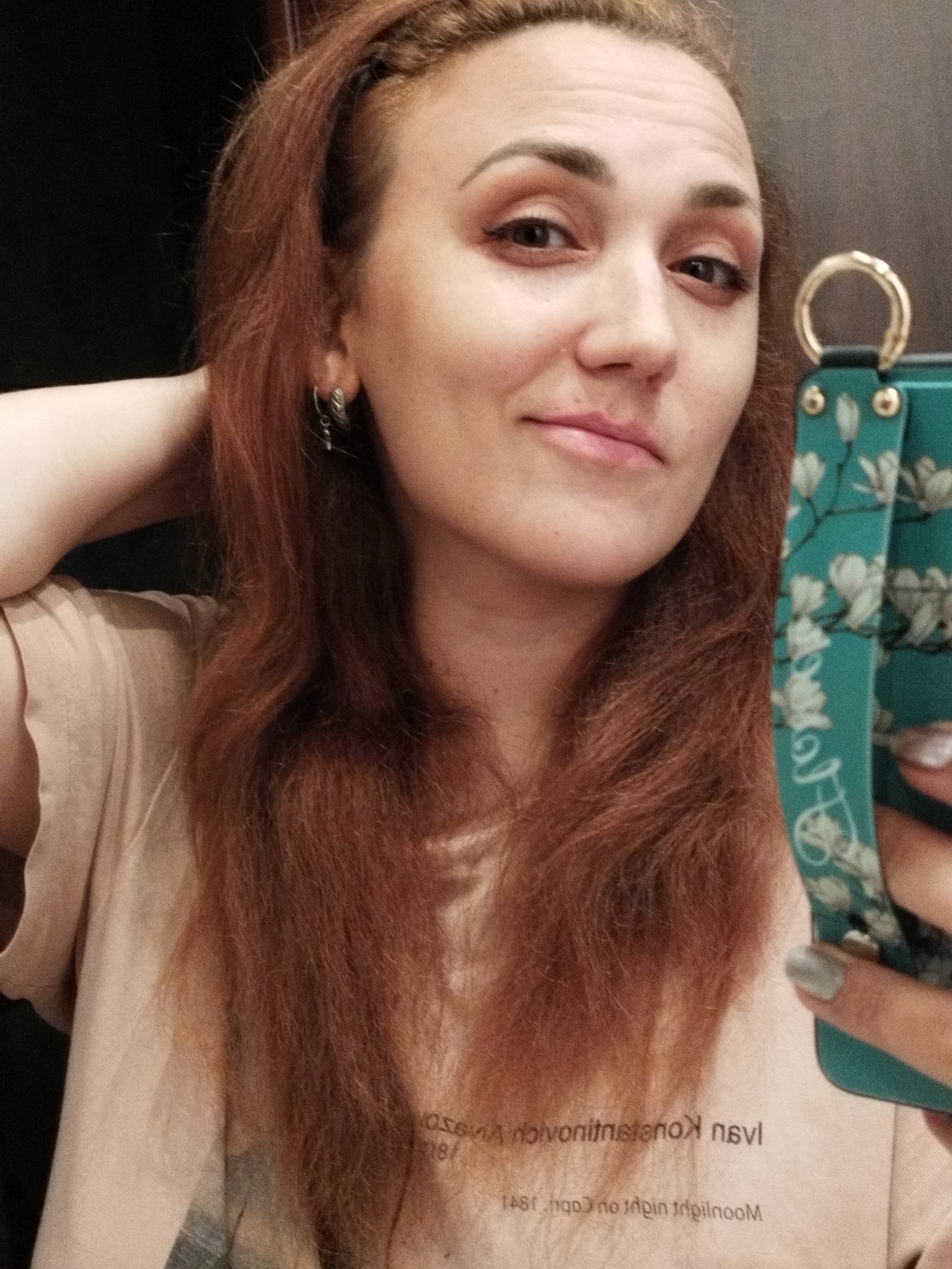 A woman with auburn hair is taking a selfie in what appears to be an indoor setting with a dark background. She is wearing a light-colored t-shirt with some text visible on it. She is holding a phone with a turquoise case adorned with a floral pattern. Her right hand is touching her hair, and her left hand is holding the phone. She is looking at the camera with a slight smile. The image is taken from the chest up.