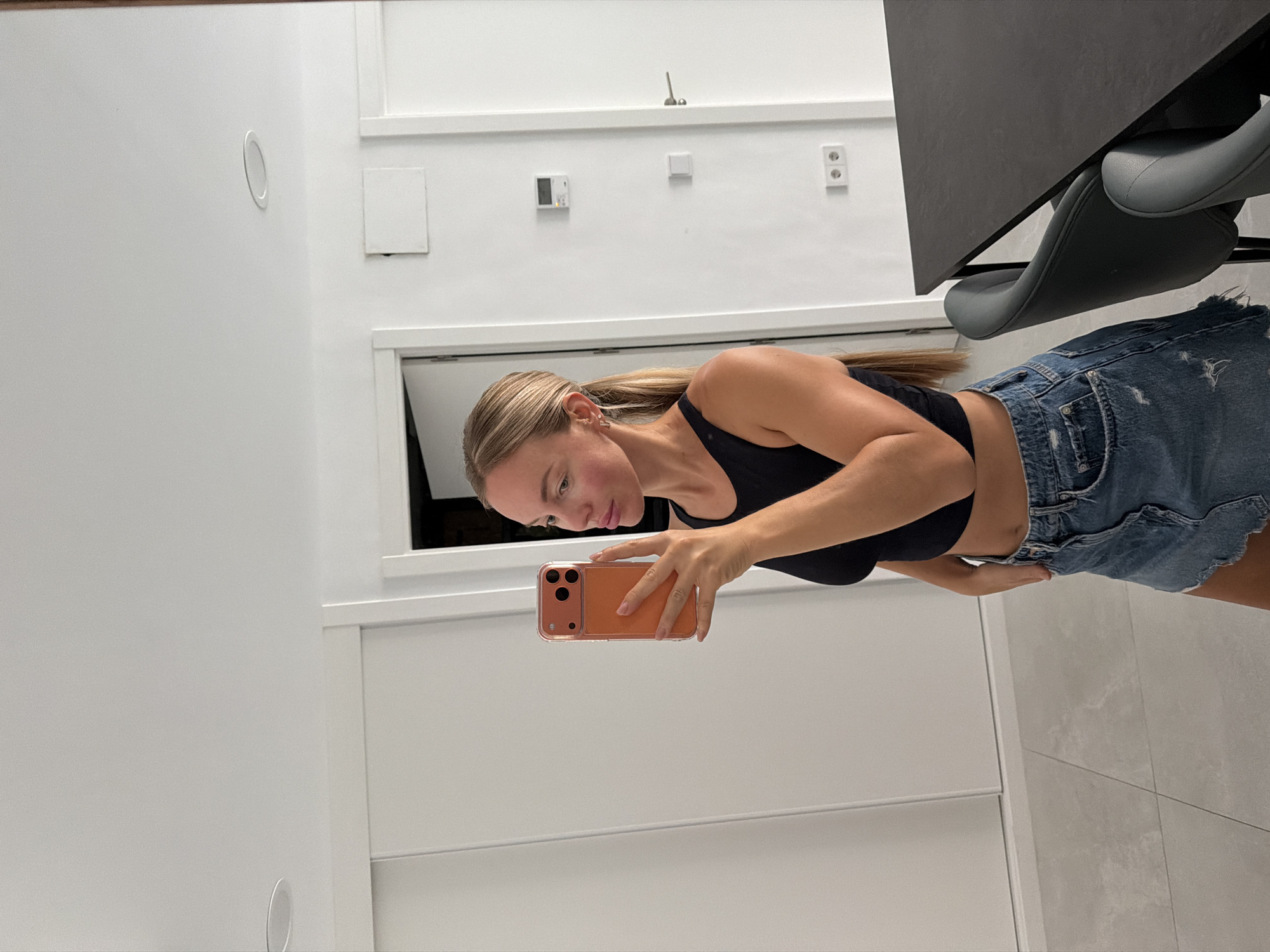 A young woman with blonde hair pulled back in a ponytail is taking a mirror selfie in a modern apartment in Finestrat, Spain. She is wearing a black crop top and a distressed denim mini skirt. The background shows a hallway with white doors, modern lighting fixtures, and electrical outlets on the wall. To the right, a black table with gray chairs is visible. The woman is holding an orange-cased smartphone to take the picture.