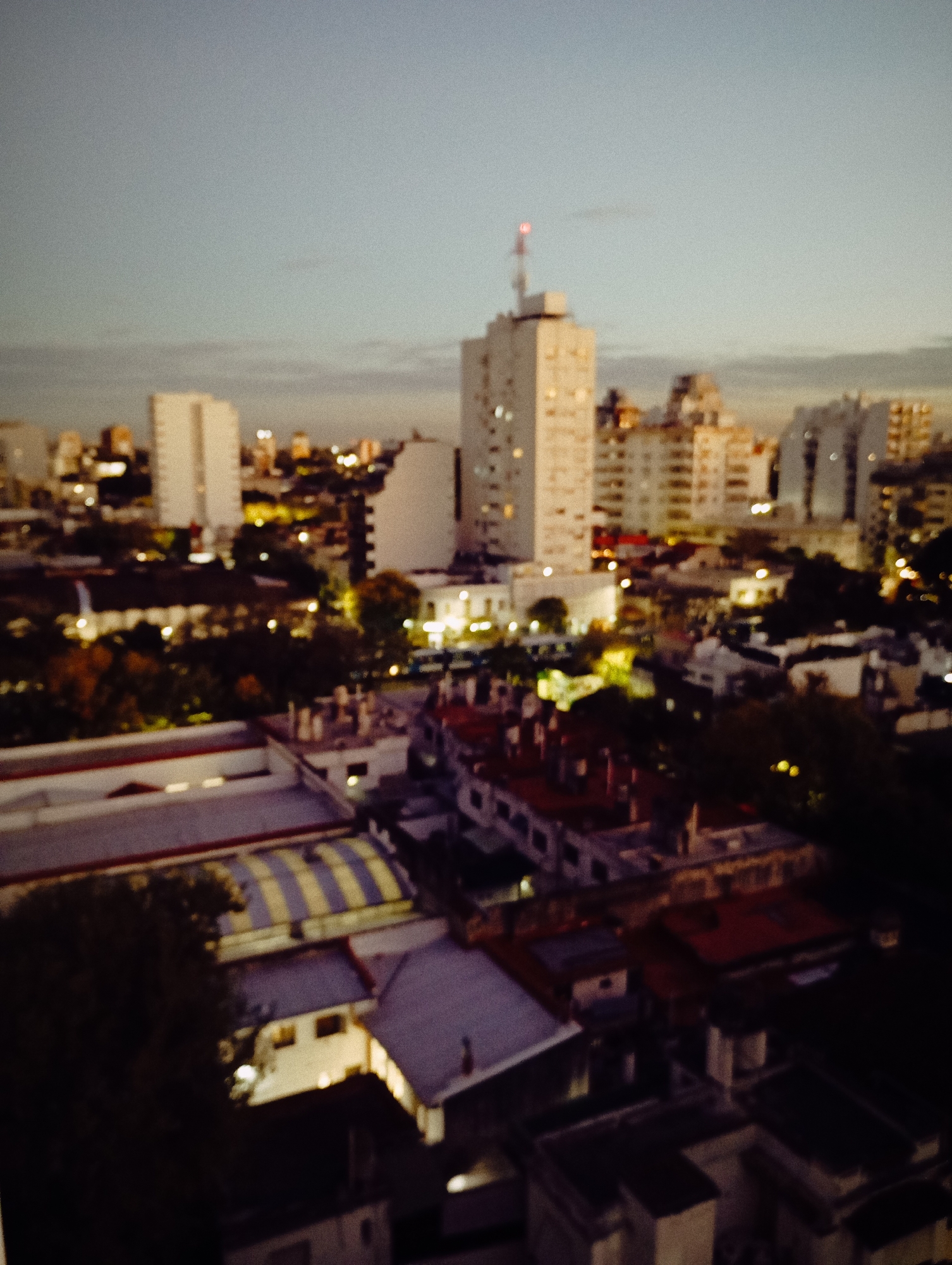 The image shows a nighttime cityscape, likely taken from an elevated position, possibly a high-rise building. The scene is softly lit, with a dominant tower building in the center-right. The red light is seen on top of the tower. Other buildings of varying heights are scattered around, illuminated by streetlights and building lights. Below the tower, there are residential buildings with red roofs. There are few trees visible. The scene is out of focus. Given the architectural style and urban layout, it is highly plausible that the location is Buenos Aires, Argentina.