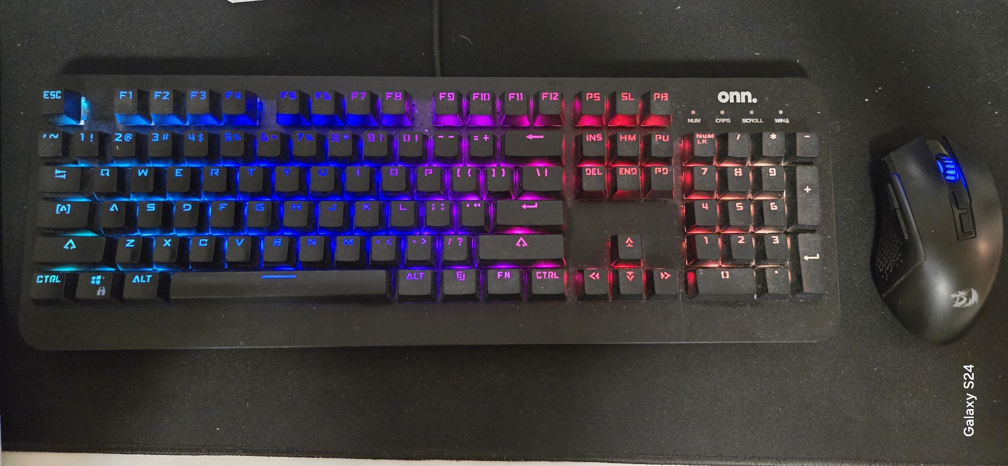 A top-down shot captures a black computer keyboard and a black gaming mouse resting on a dark fabric mouse pad. The keyboard, branded "onn.", features vibrant RGB backlighting, transitioning from blue on the left side (including the ESC and F1-F4 keys), to purple in the central section (F5-F12, alphanumeric keys), and red on the right-most sections (navigation keys, numeric keypad). Keys such as CTRL, ALT, Shift, Tab, Caps Lock, Enter, and Backspace are clearly visible, along with all alphanumeric keys, function keys (F1-F12), and media/control keys like INS, DEL, HOME, END, PG UP, PG DN, PRT SC, SCR LK, and PAUSE BRK. Indicator lights for NUM, CAPS, and SCROLL lock are present above the numeric keypad. Dust and small particles are visible on and around the keyboard keys, indicating use.
To the right of the keyboard, a black gaming mouse, bearing a faint Redragon logo on its side, displays blue LED lighting on its scroll wheel and DPI buttons. The mouse is positioned to the right of the keyboard, as if in active use. The mouse pad is dark gray or black. Along the bottom right edge of the mouse pad, the text "Galaxy S24" is visible vertically, suggesting the device used to take the photograph. The scene depicts an indoor, static setup with no direct activity taking place.