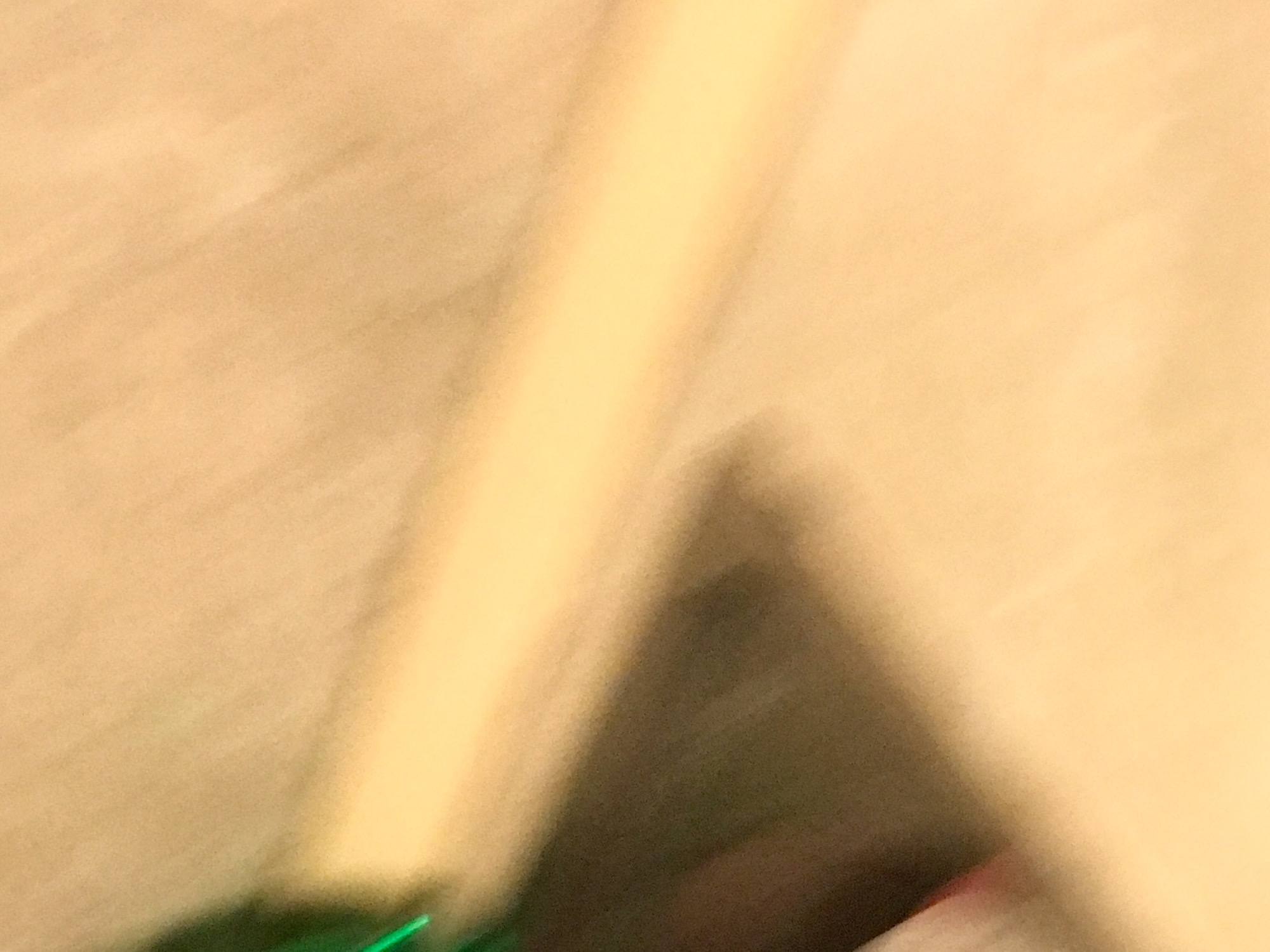 The image is a blurry, close-up shot with an indeterminate background of light brown and tan. A dark, triangular or pyramidal shape with undefined edges is visible at the bottom center, partially obscuring what might be the background. A strip of light yellow is oriented vertically. A hint of green is visible in the lower left corner, along with a smudge of red towards the lower right. The scene is too indistinct to identify a specific location in Yerevan, Armenia.