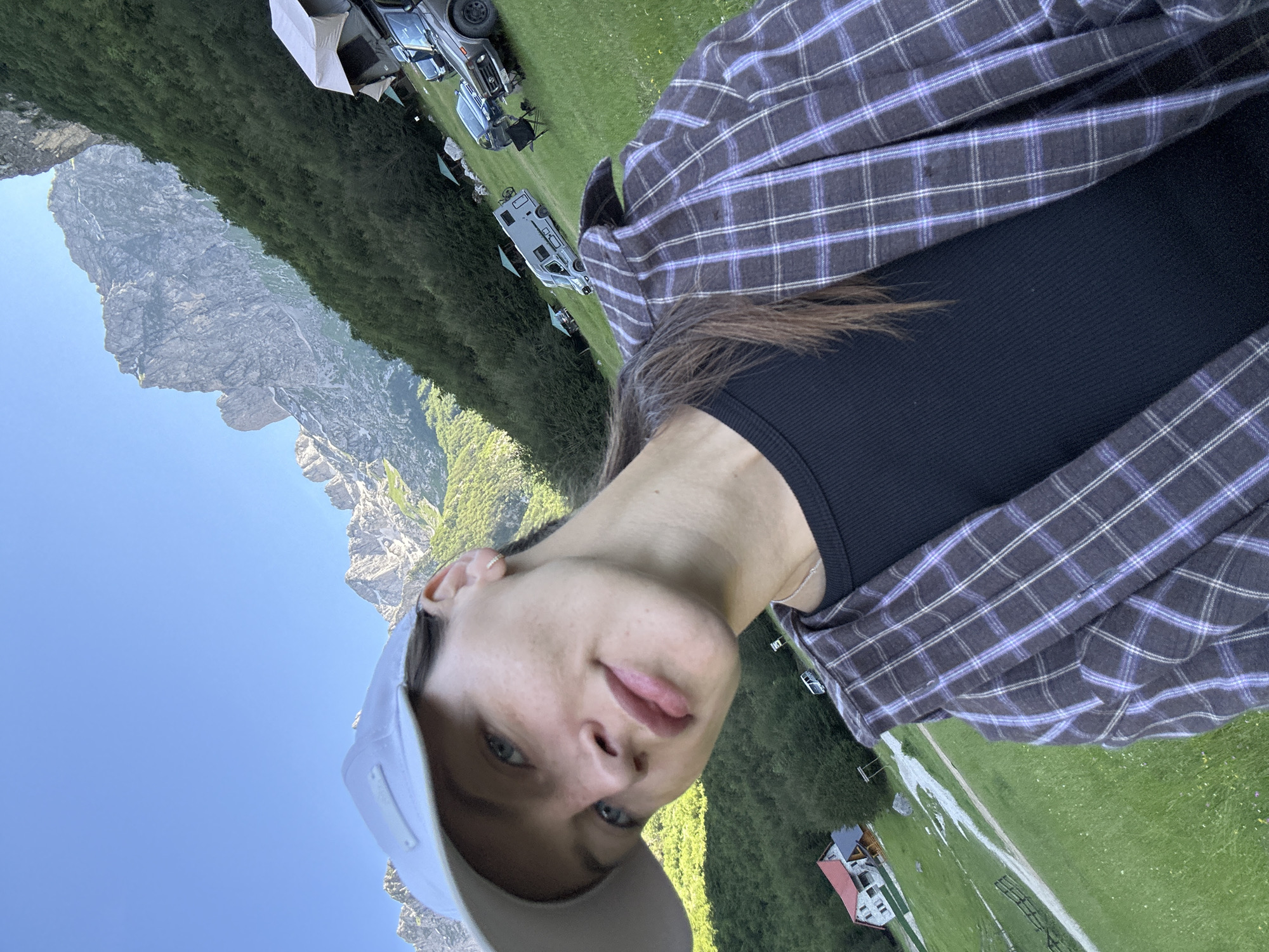 In the city of Dolja, Montenegro, a young woman with blue eyes, wearing a white baseball cap and a plaid shirt over a black top, smiles at the camera. She is positioned in the foreground, with a backdrop of lush green mountains and a clear blue sky. Several camper vans and tents are visible on the grassy terrain, suggesting a camping or outdoor recreational area. A stone house with a red roof is situated in the lower left corner of the frame. The overall scene conveys a sense of outdoor adventure and natural beauty.